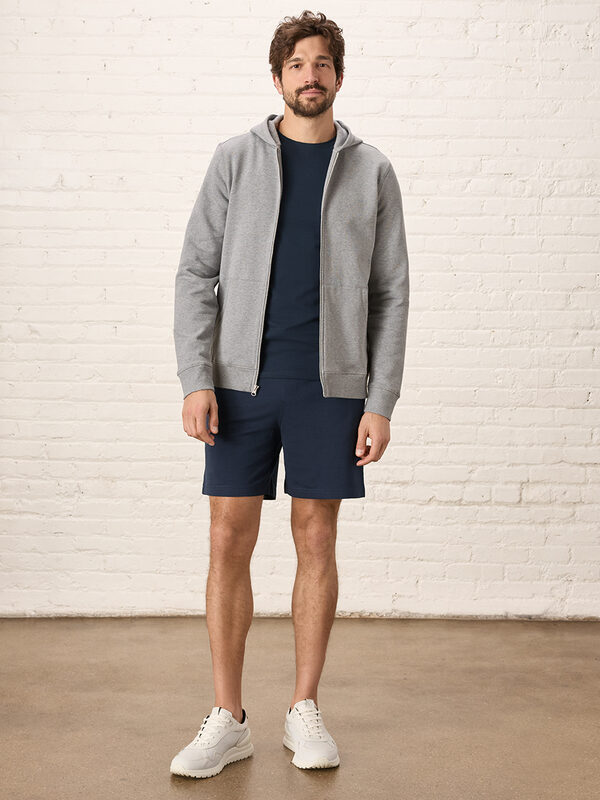 A man stands indoors against a white brick wall, wearing a grey zip-up hoodie, a navy T-shirt, navy shorts, and white sneakers.
