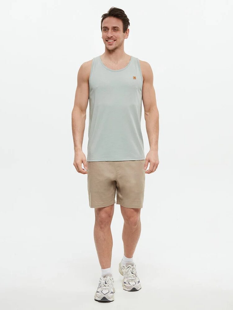 Man standing against a plain background wearing a light blue tank top, beige shorts, and white sneakers.
