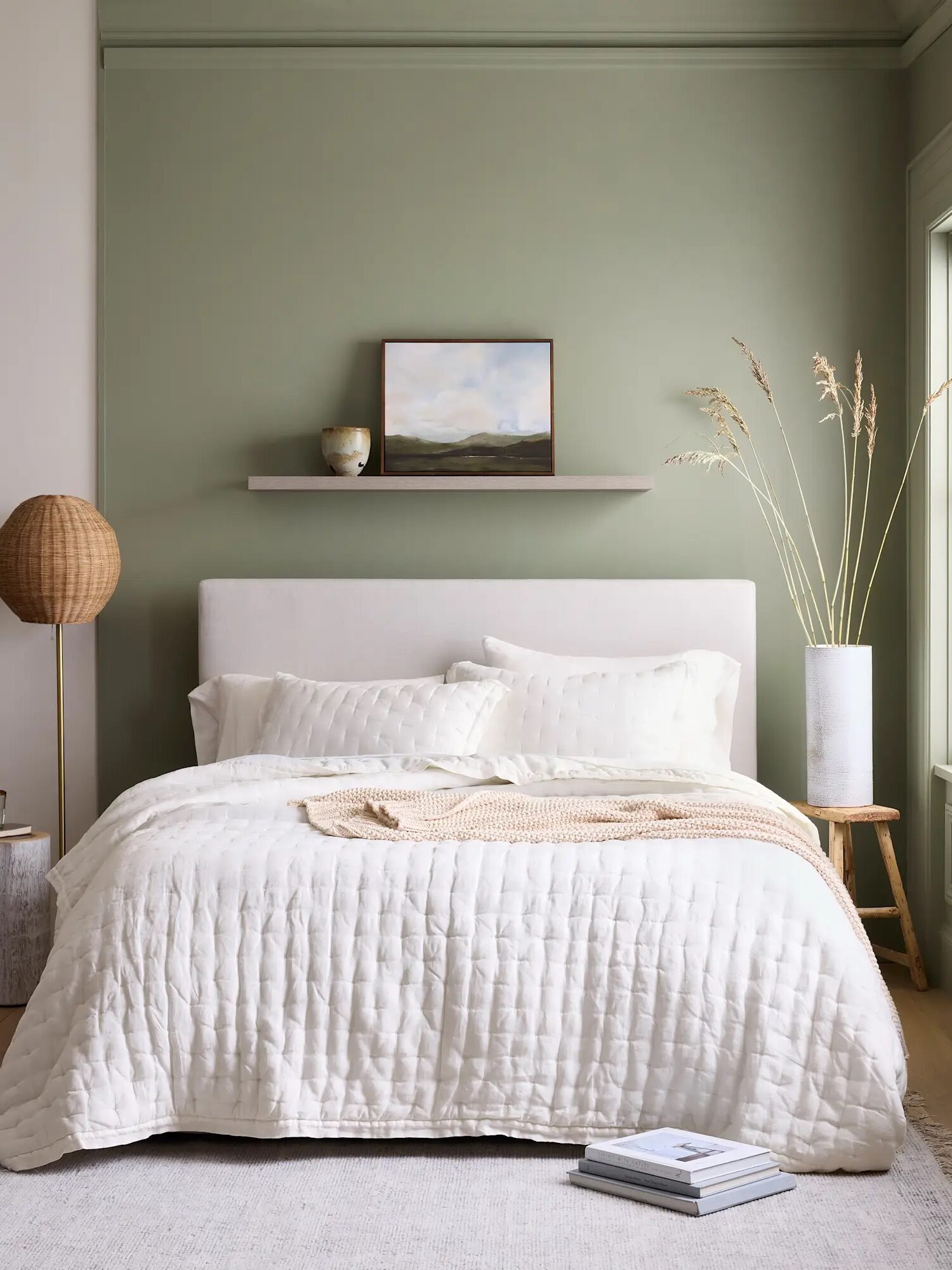 A neatly made bed with a cream quilt, pillows, and throw sits against a sage green wall with a shelf holding a painting and decor; a round lamp and tall vase are on either side.