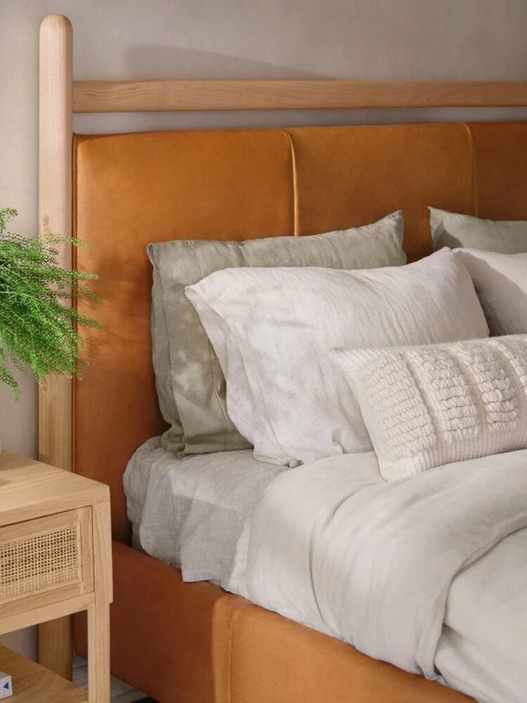 A neatly made bed with beige and sage bedding, an orange upholstered headboard, a wooden nightstand, and a green potted plant.