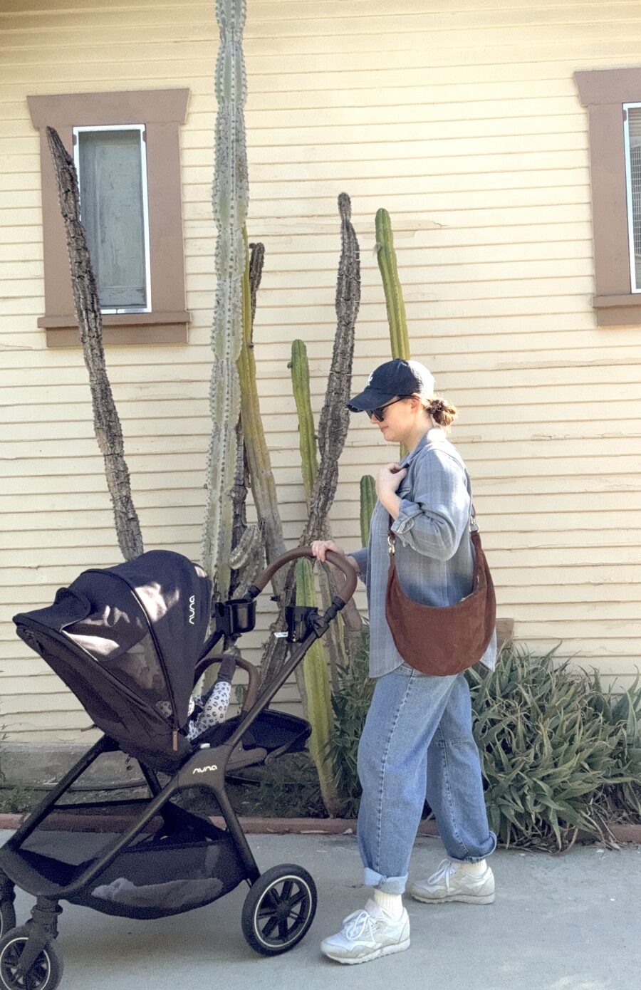 A woman in casual clothes and a baseball cap pushes a stroller with a baby past tall cacti in front of a beige house with two windows.