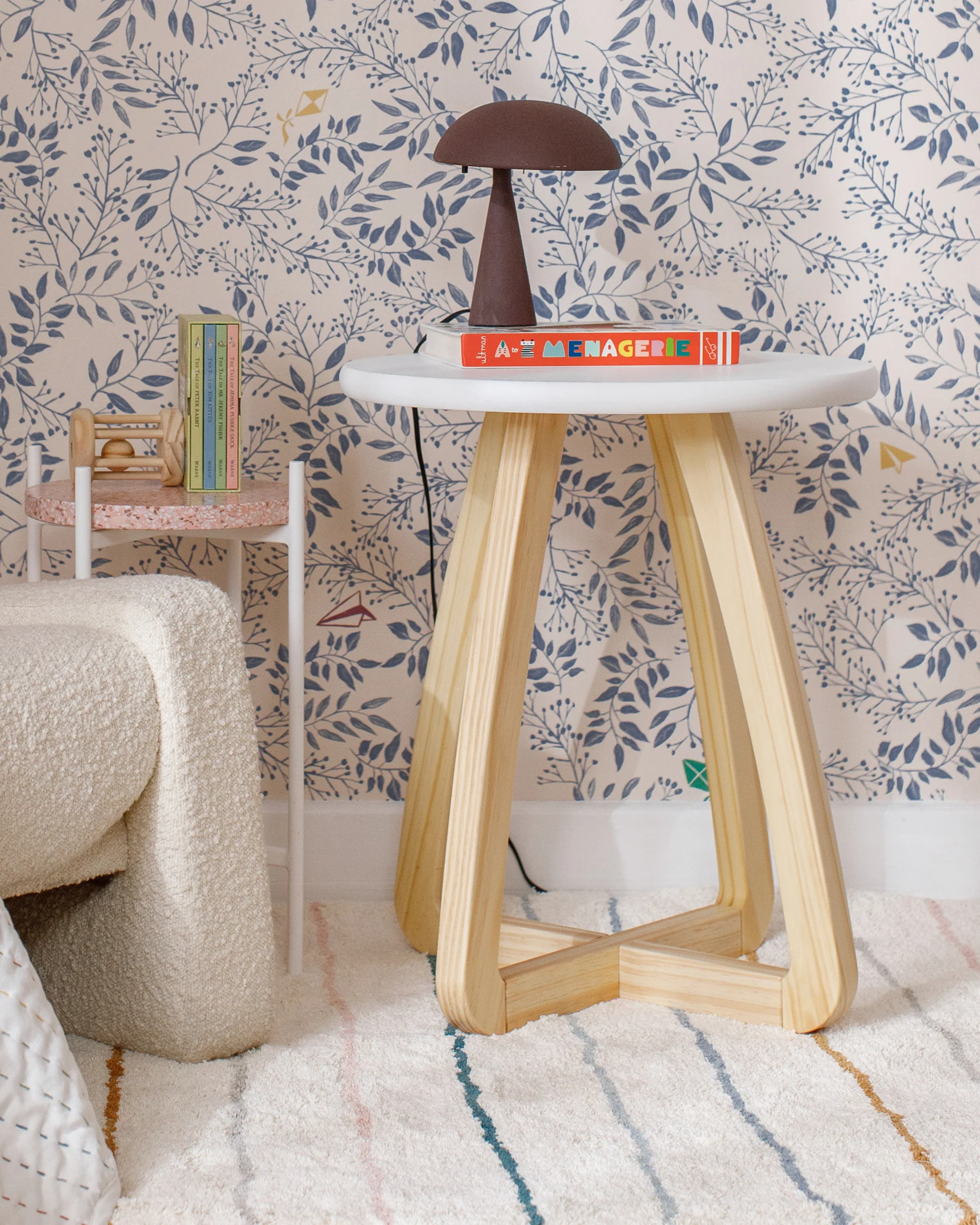 A small wooden side table holds a brown lamp and books, next to a patterned wall, with another table, books, and a white chair nearby.