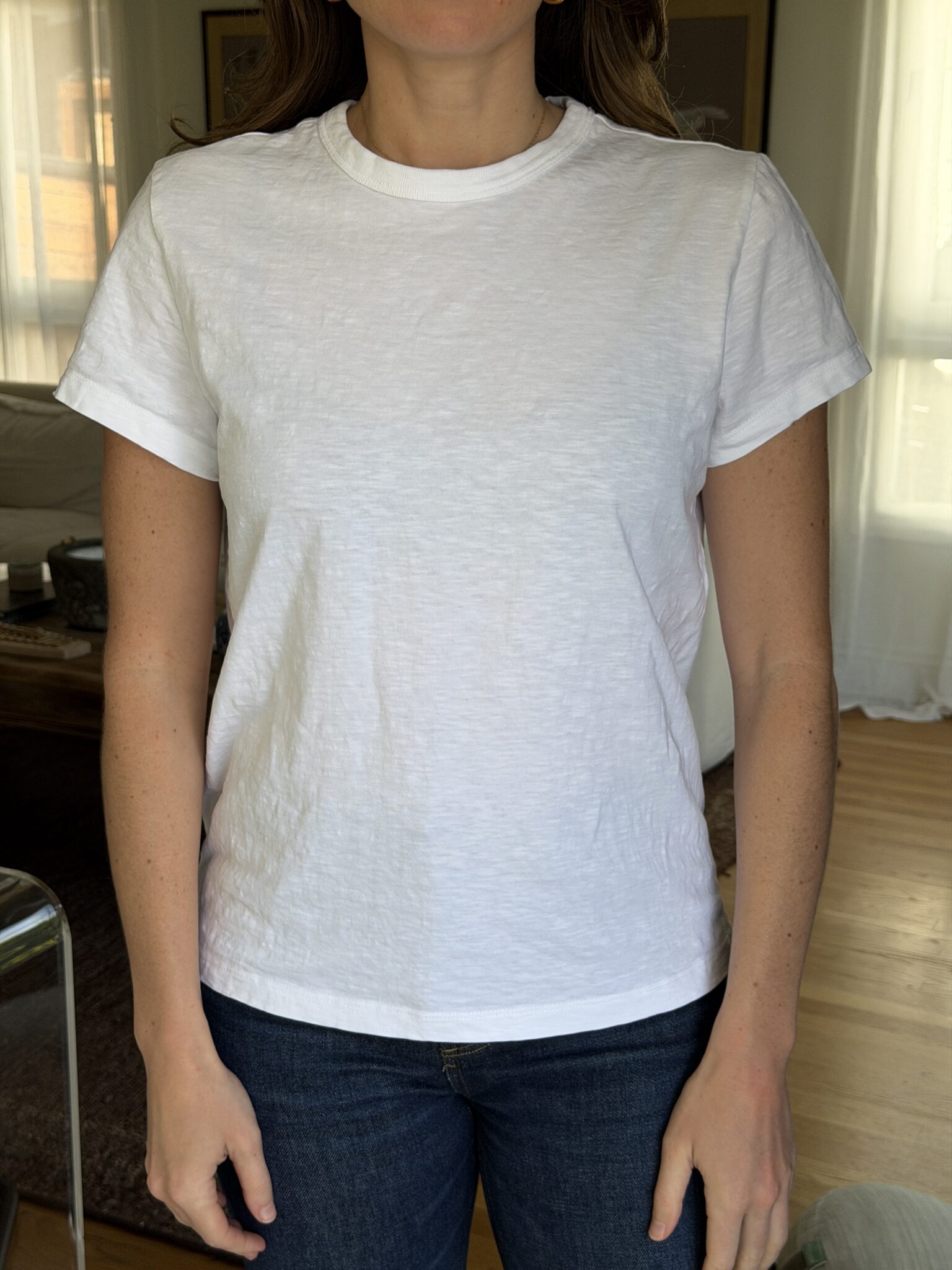 A person wearing a plain white short-sleeve t-shirt and dark jeans stands indoors in front of a window with light curtains.