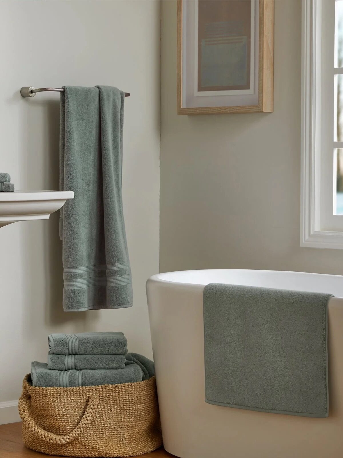 A bathroom with a white tub, a sage green organic towel draped over it, a matching towel on a rack, folded organic towels in a woven basket, and a framed picture on the wall.