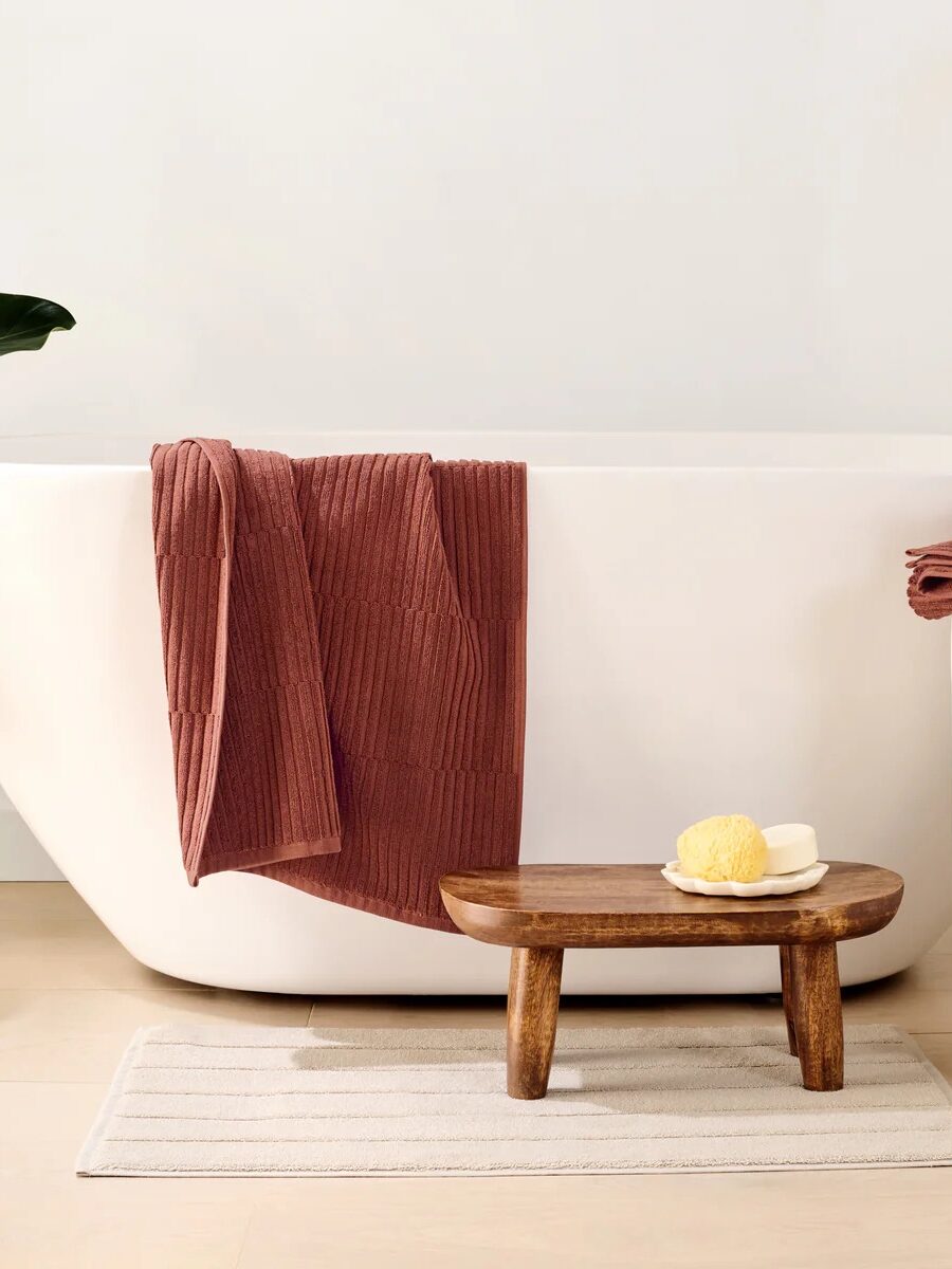A modern bathroom with a white freestanding tub, organic rust-colored towels, a small wooden stool holding a sponge and soap, and a beige mat on light wood flooring.