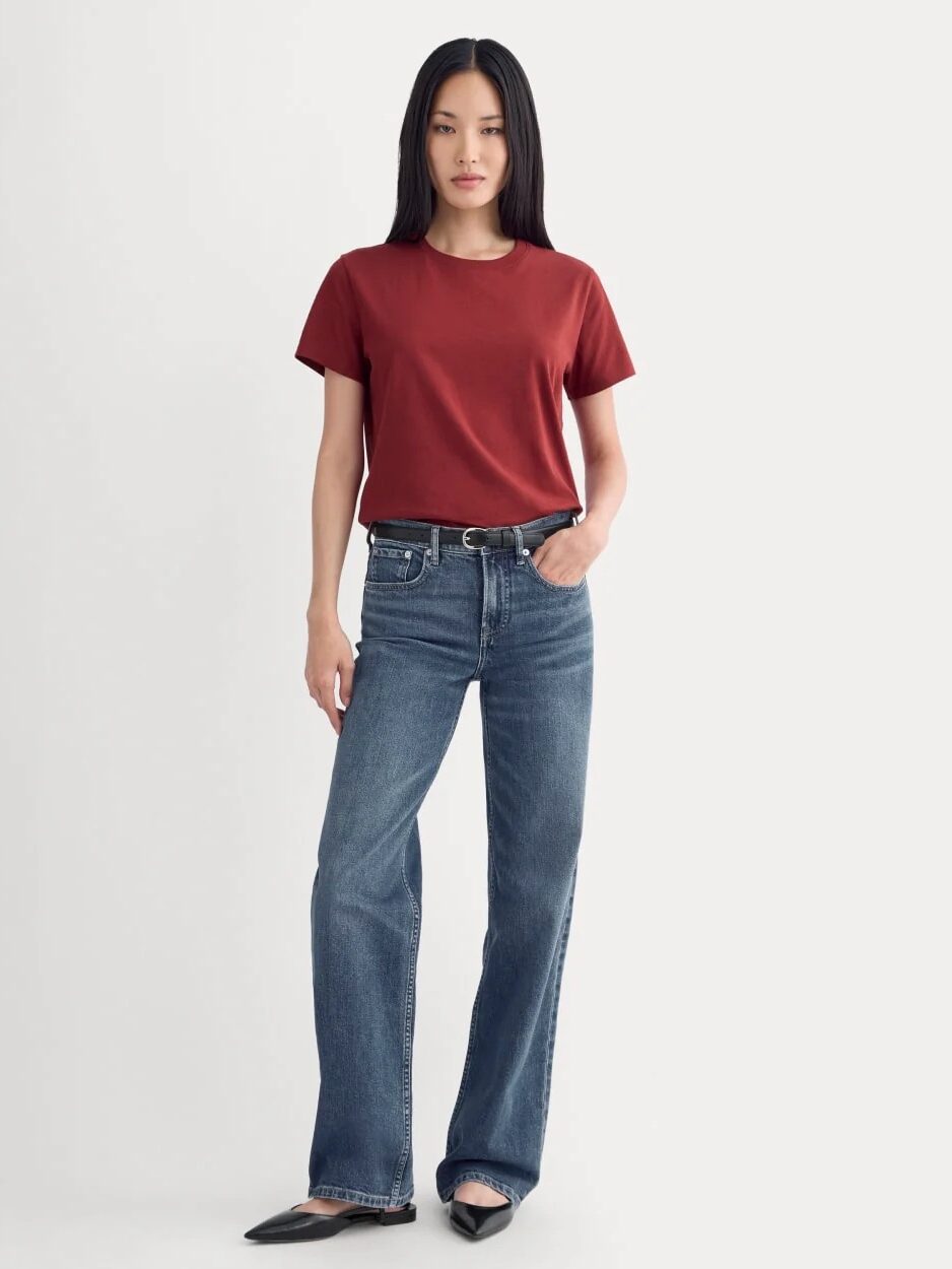 A woman stands facing forward, wearing a red short-sleeve t-shirt, blue jeans, and black shoes against a plain light background.
