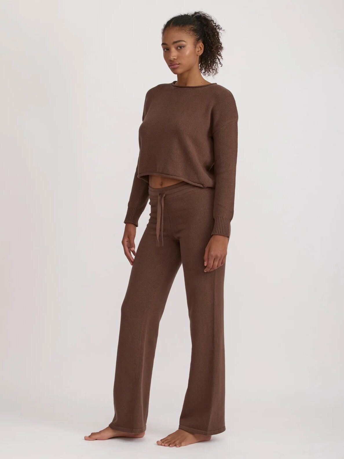A woman stands barefoot against a plain background, wearing a matching brown knit long-sleeve top and drawstring pants set.