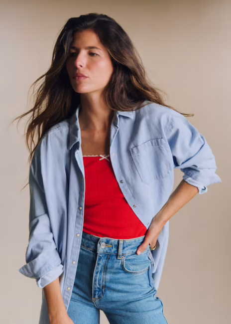 Woman wearing a light blue button-up shirt over a red top and blue jeans, standing with one hand on her hip against a plain background.