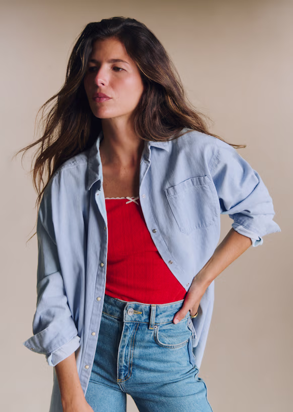 Woman wearing a light blue button-up shirt over a red top and blue jeans, standing with one hand on her hip against a plain background.