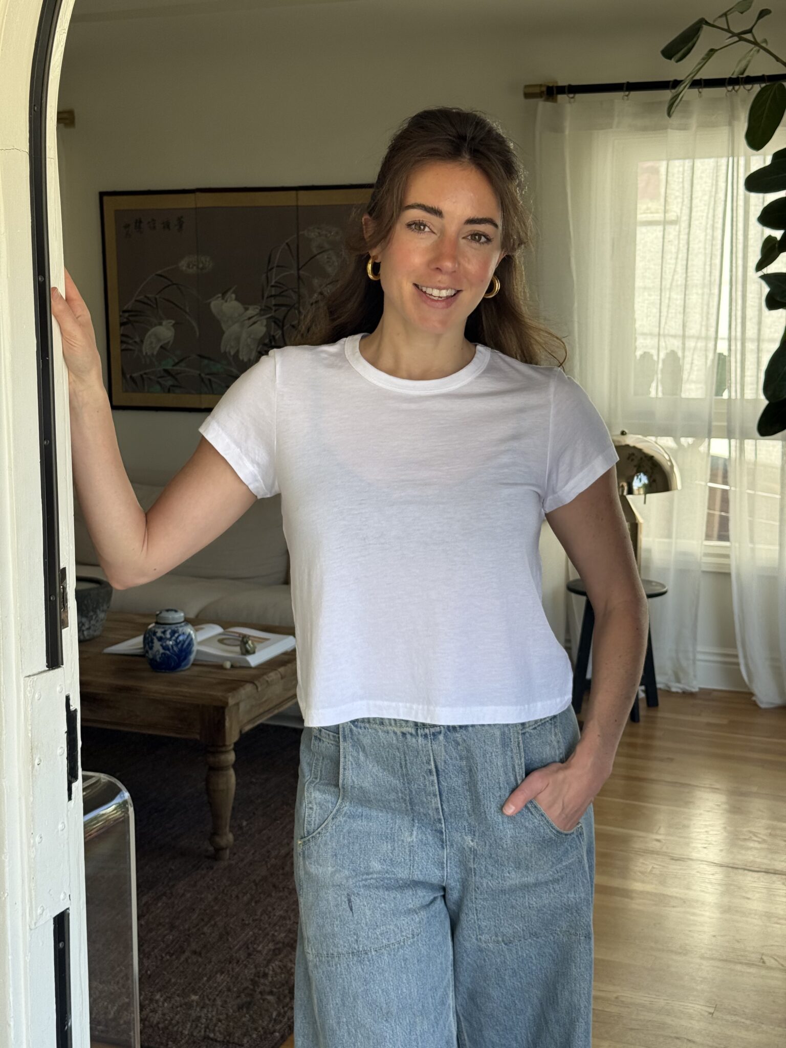 A woman stands indoors by a doorway, wearing a white t-shirt and high-waisted jeans, with one hand in her pocket and the other resting on the doorframe.