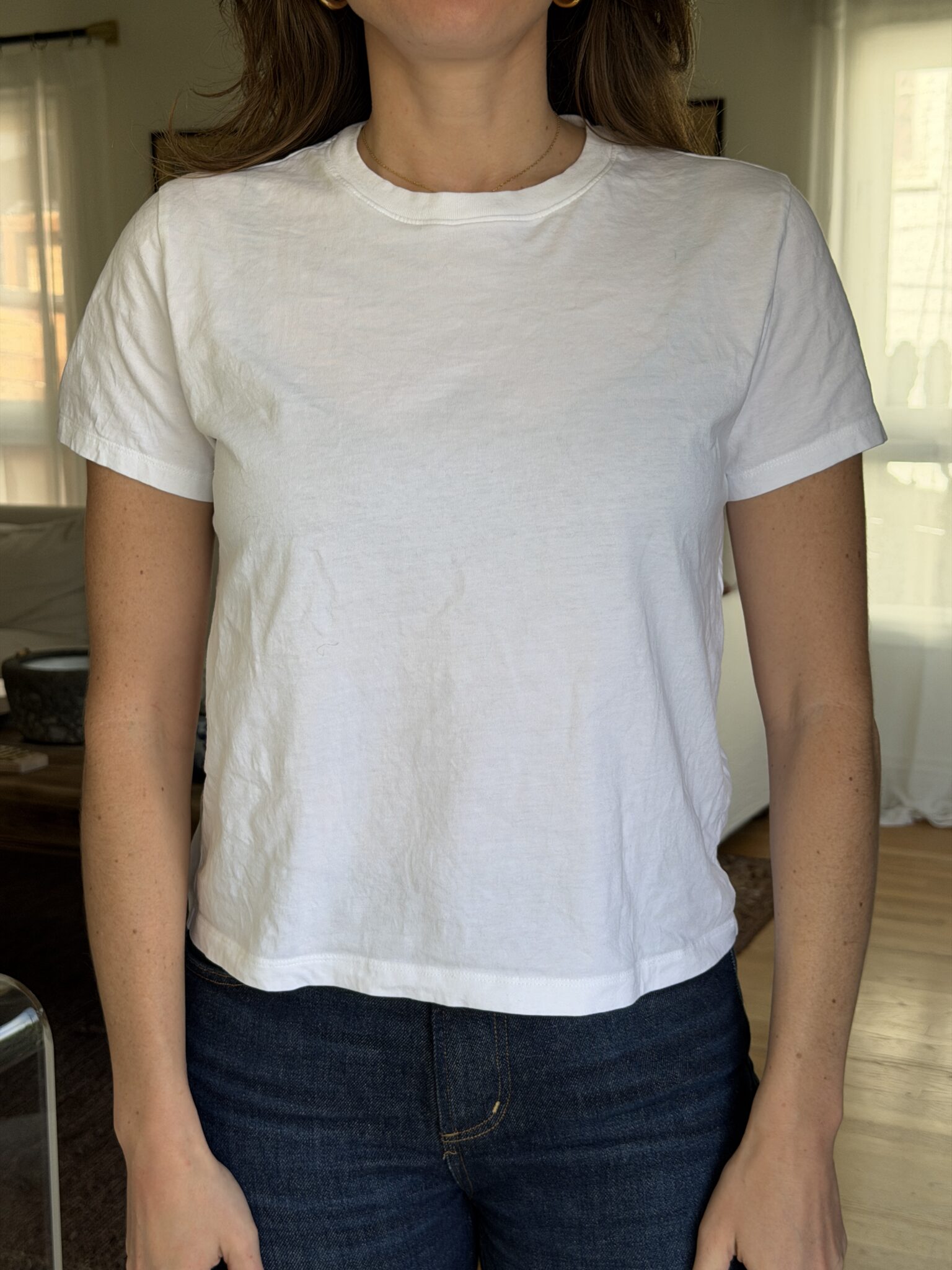 A person standing indoors wearing a plain white short-sleeve t-shirt and dark jeans.