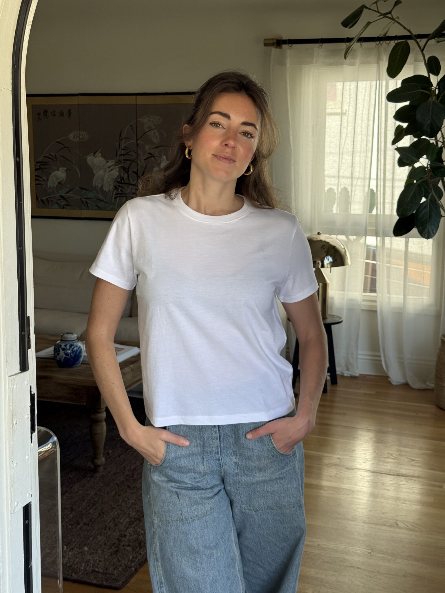 Woman standing indoors with hands in jean pockets, wearing a white t-shirt, in a well-lit living room with plants and curtains in the background.