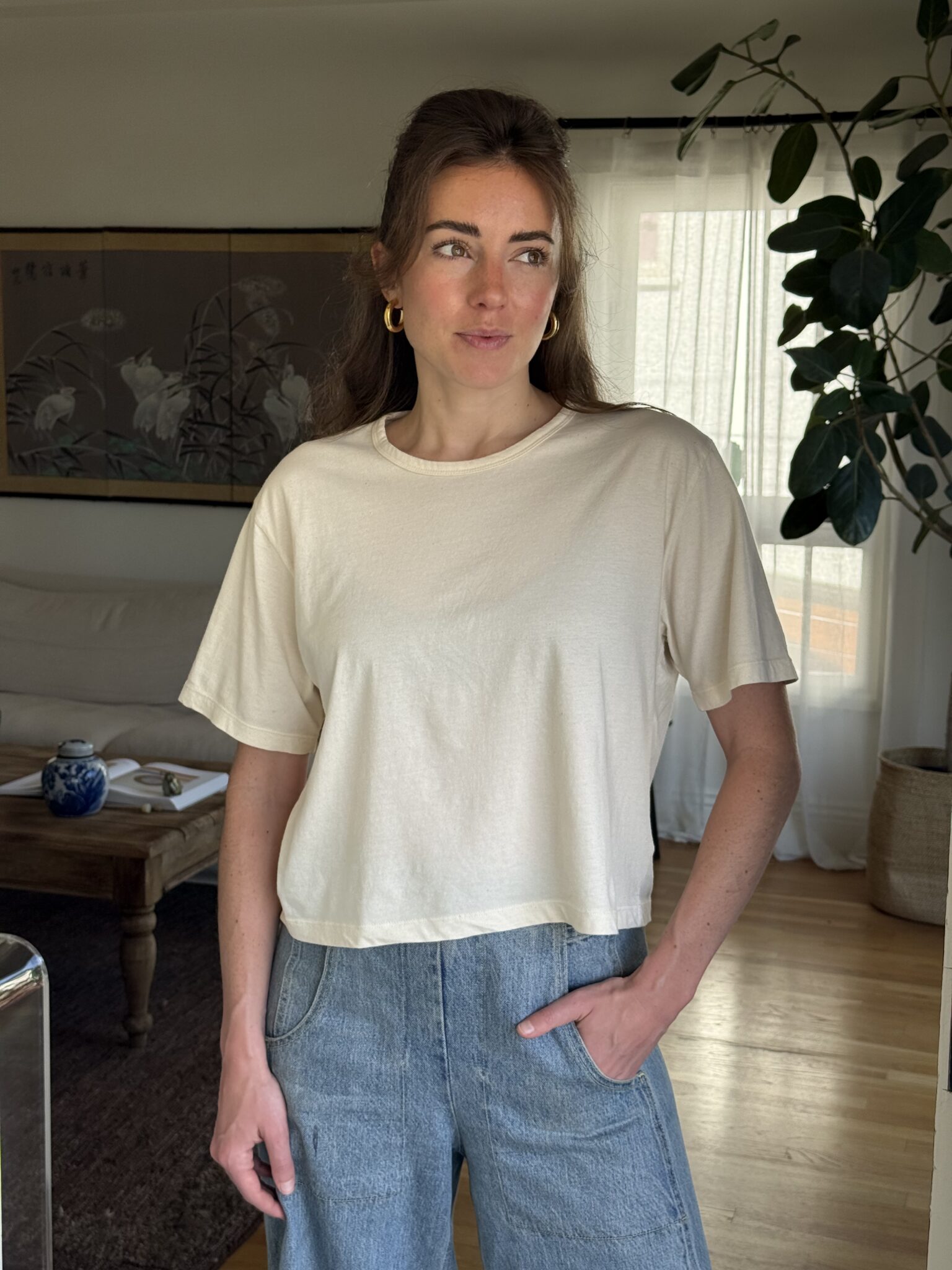 A woman with long brown hair wearing a beige t-shirt and blue jeans stands indoors with one hand in her pocket. There is a plant and a coffee table in the background.
