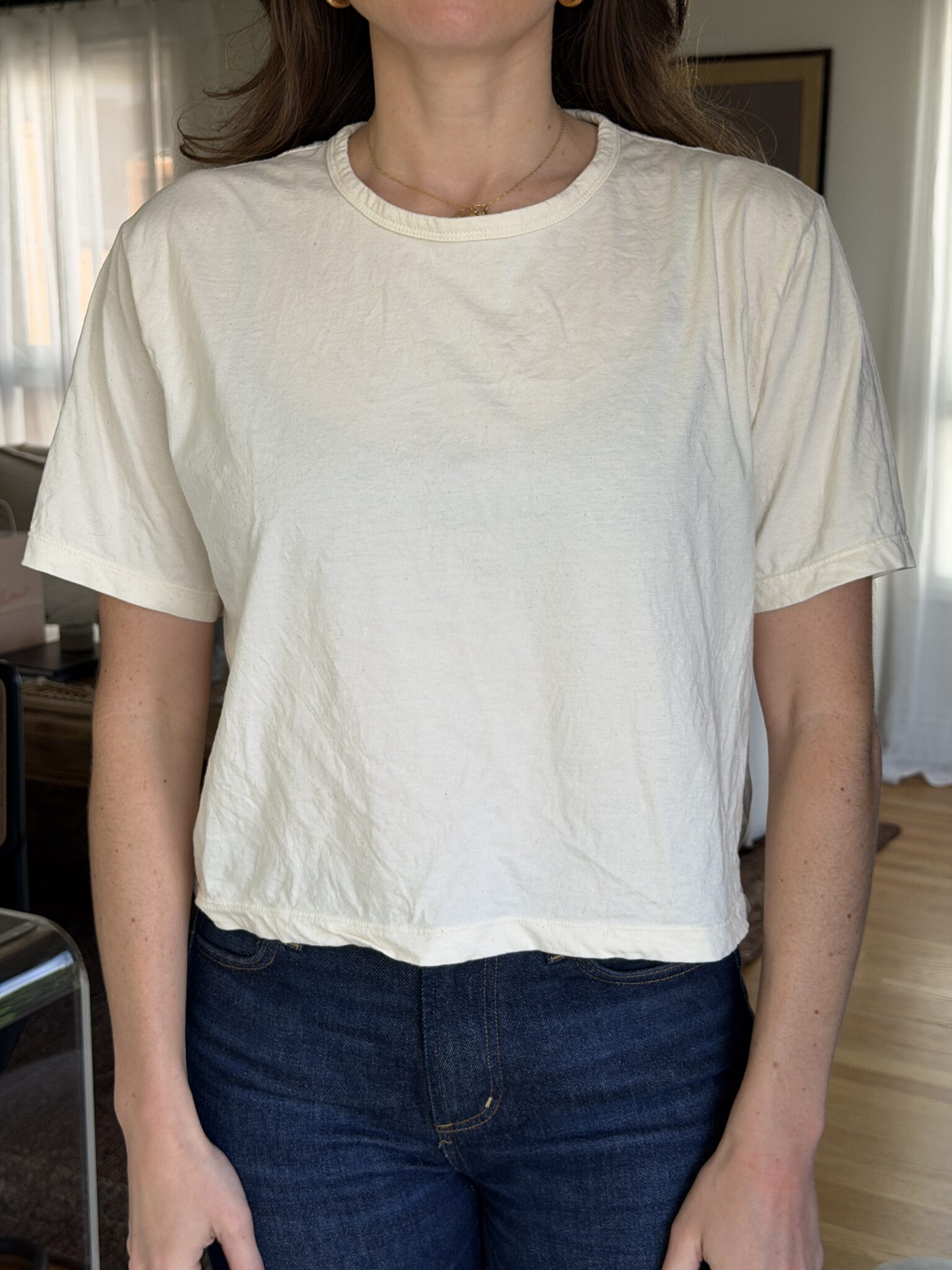 A person wearing a plain, short-sleeve, off-white T-shirt and dark blue jeans stands indoors with natural light coming through the window.