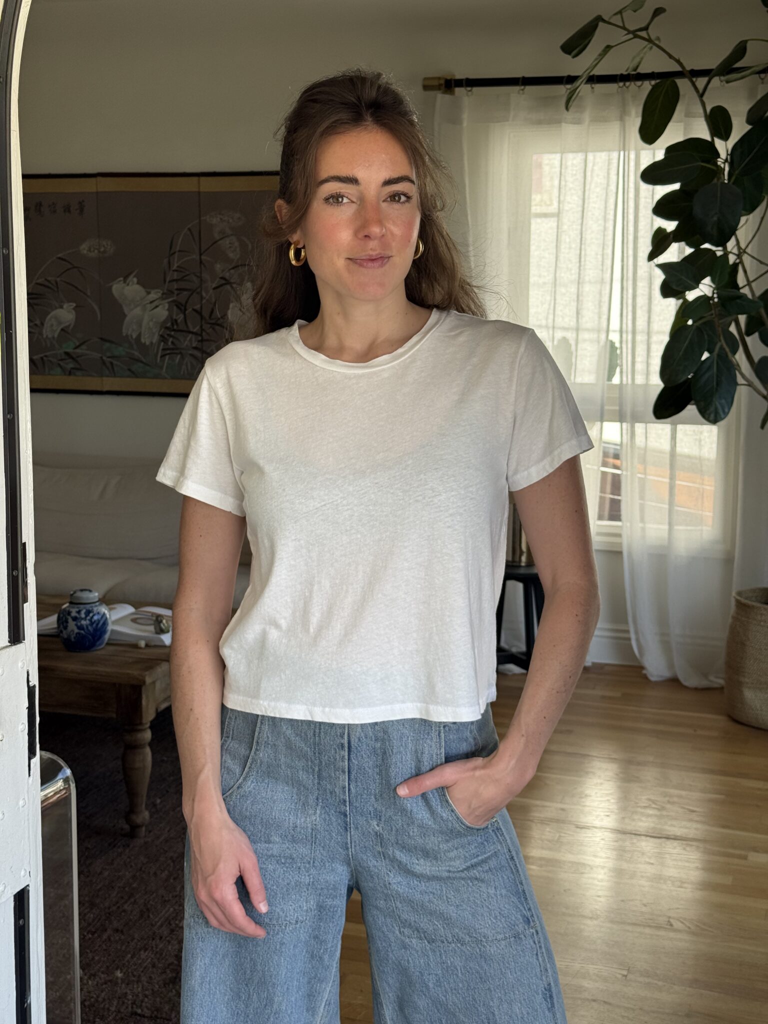A woman with long brown hair wearing a white t-shirt and blue jeans stands indoors with one hand in her pocket, in a well-lit room with plants and a wooden floor.