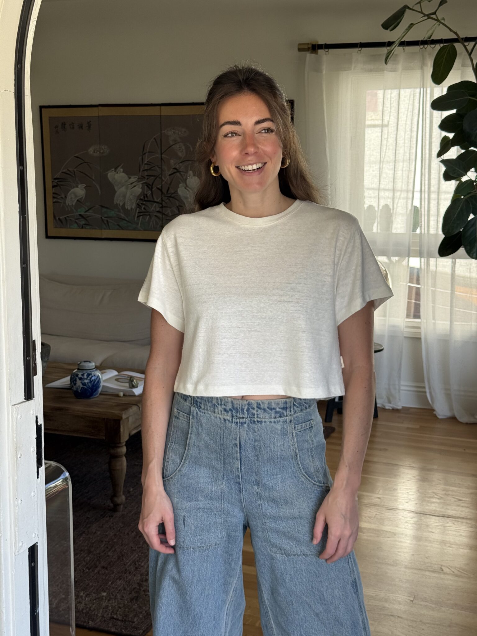 A woman stands indoors wearing a white cropped t-shirt and high-waisted blue jeans, smiling, with a living room and large plant in the background.