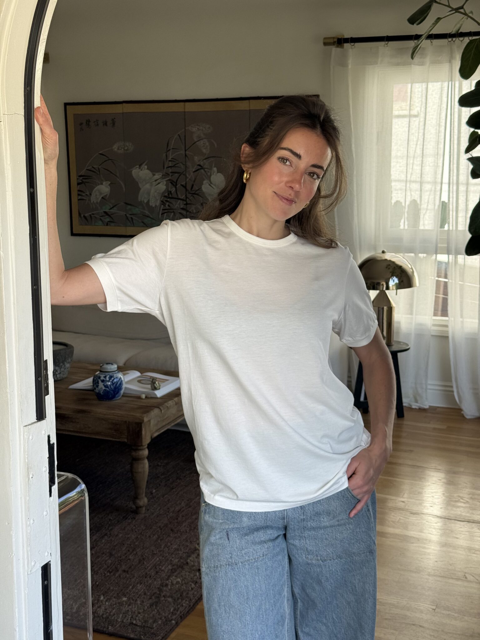 Woman wearing a white t-shirt and blue jeans stands in a living room, resting one arm on a doorway and looking at the camera. The room has a wooden table, a lamp, and framed art on the wall.