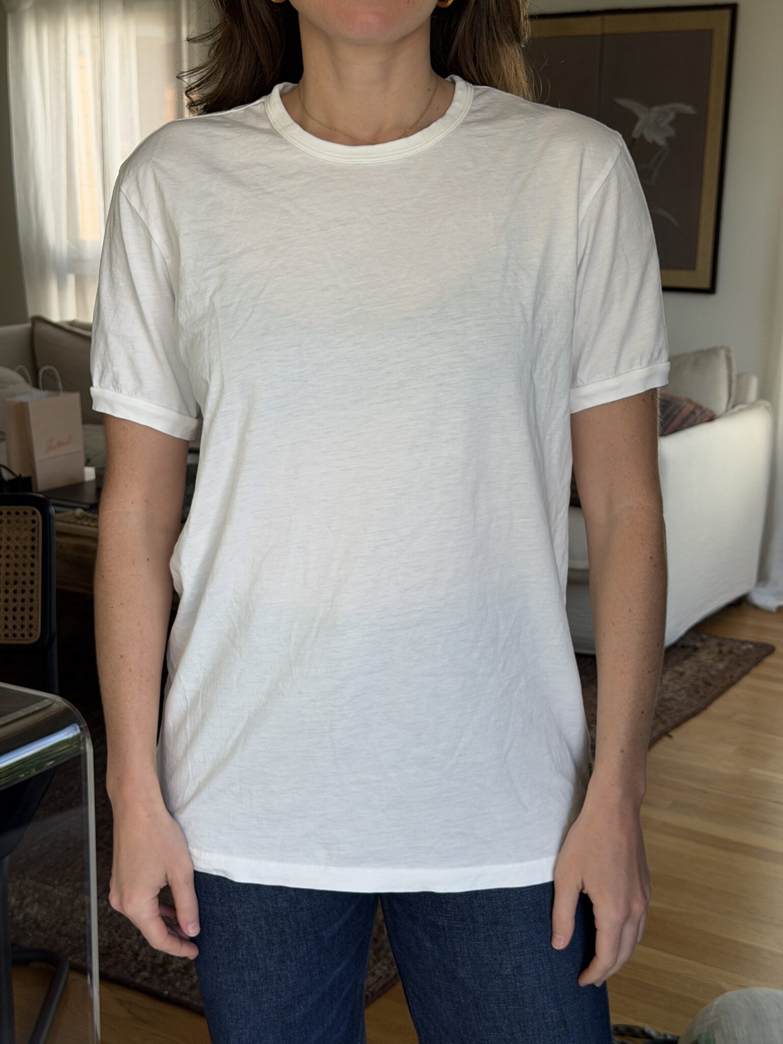 A person standing indoors wearing a plain white short-sleeve t-shirt and dark blue jeans, with their arms relaxed at their sides.