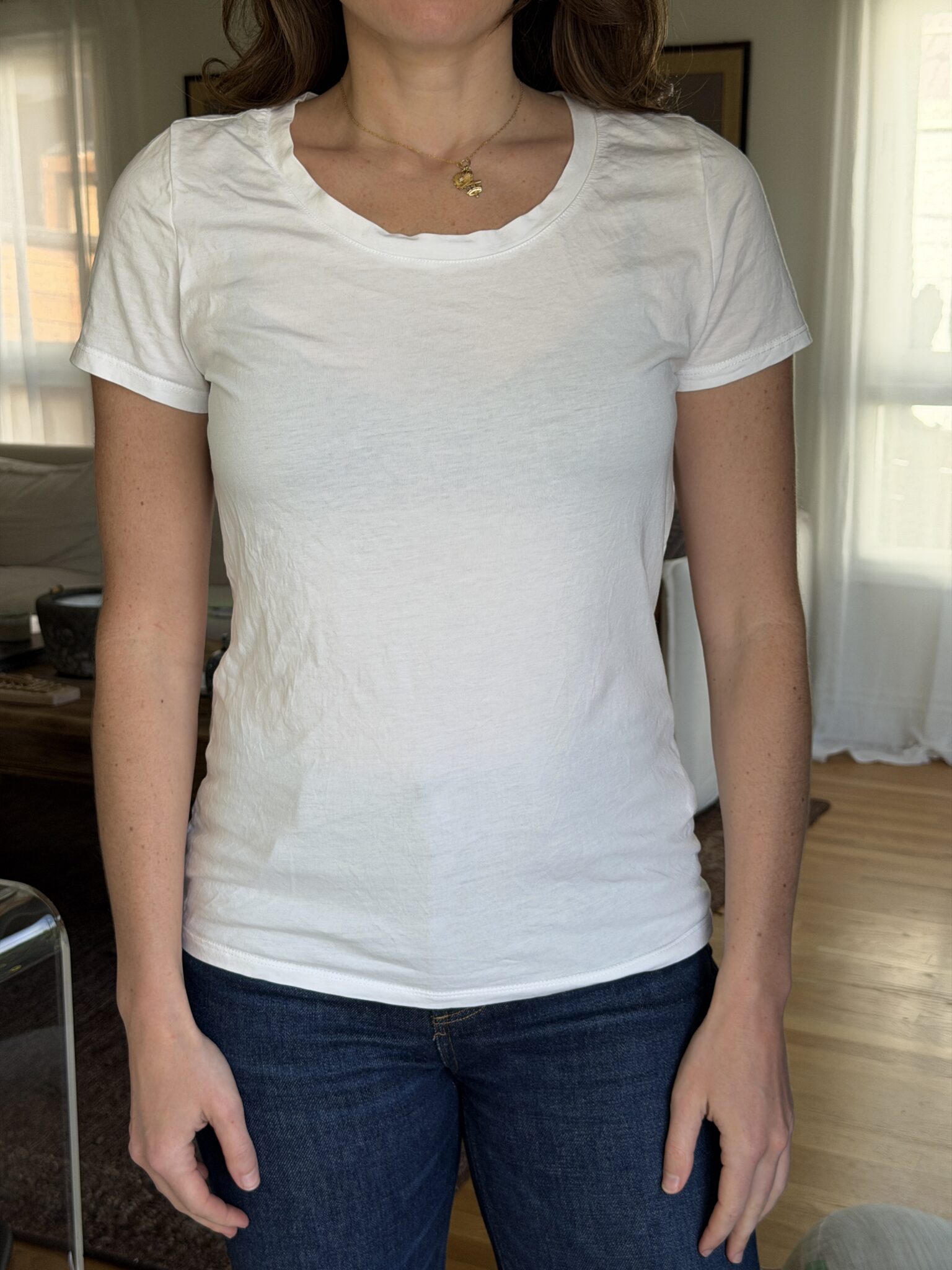 A person wearing a plain white short-sleeve t-shirt and blue jeans stands indoors with their arms relaxed at their sides.