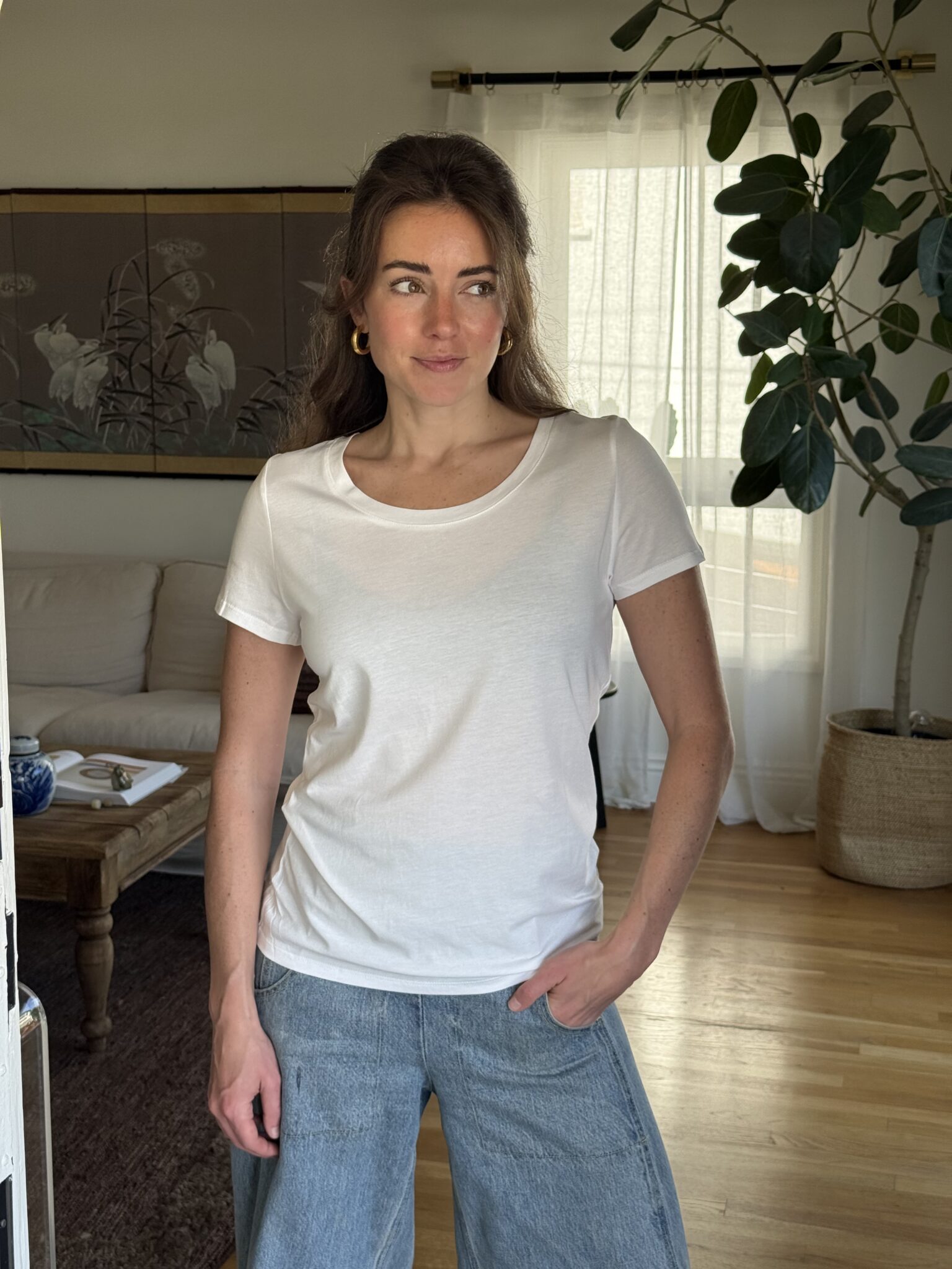 A woman stands indoors wearing a white t-shirt and blue jeans, with one hand in her pocket, in a living room with plants and a sofa in the background.