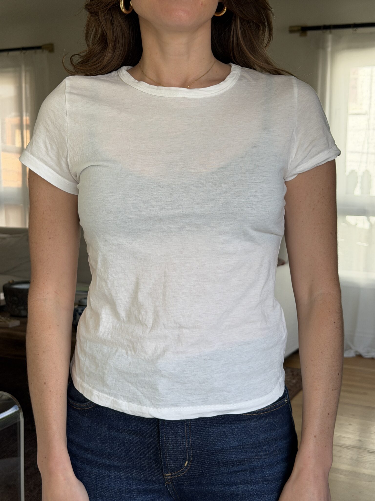 A person wearing a fitted, semi-sheer white t-shirt and dark jeans stands indoors in a well-lit room.