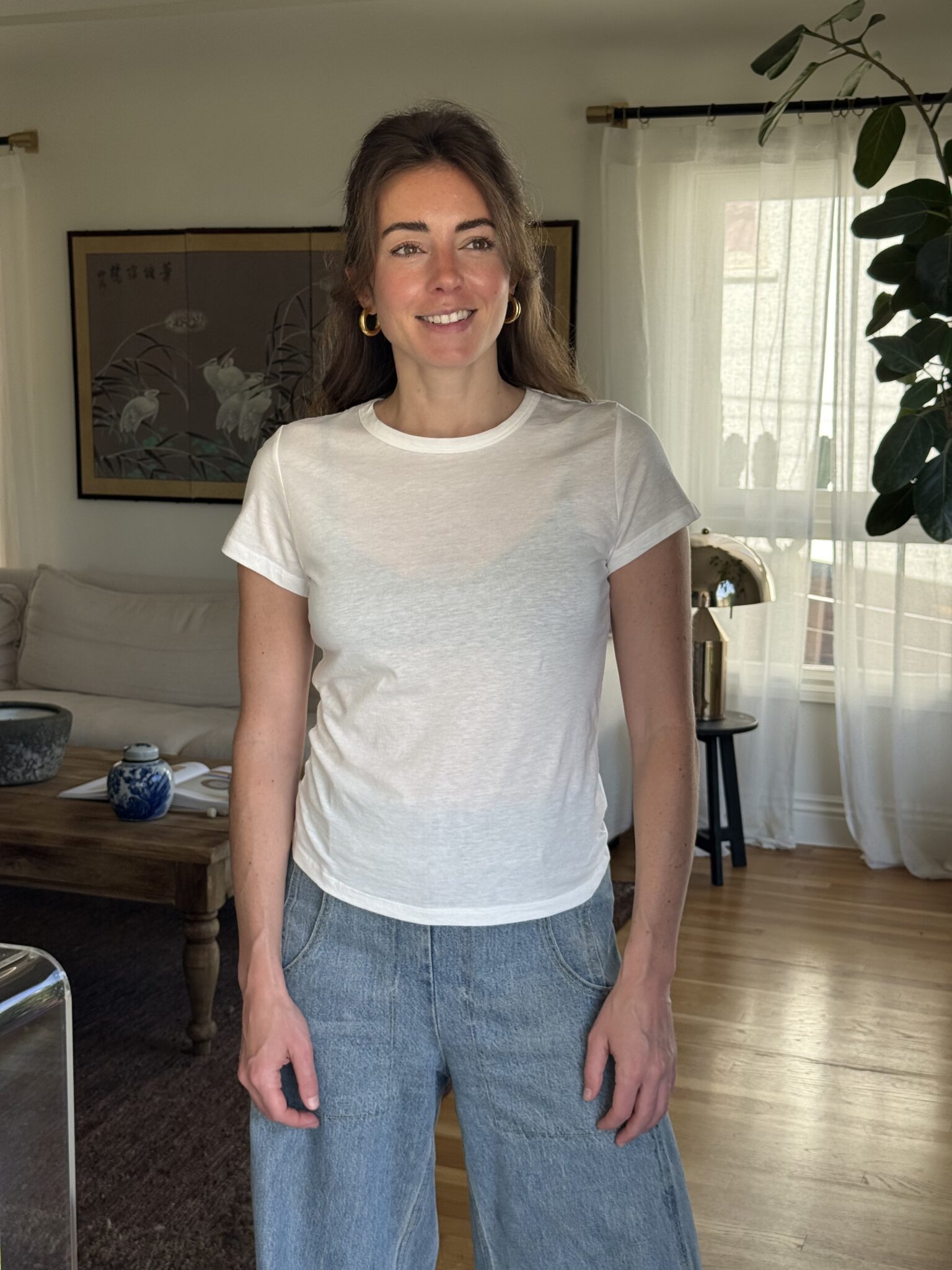 A woman stands in a living room wearing a white t-shirt and blue jeans, with light coming through sheer curtains and a potted plant nearby.