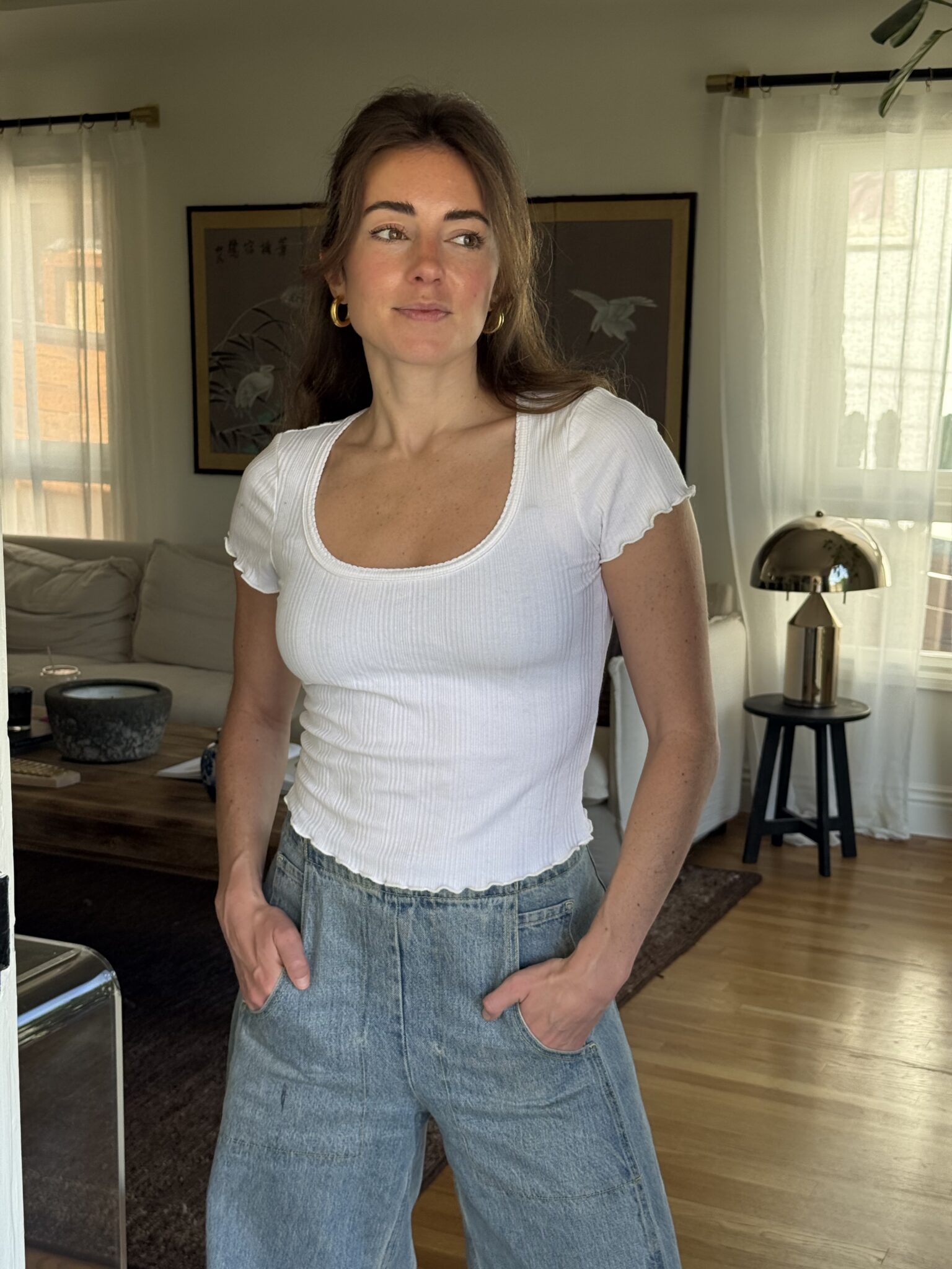 A woman in a white fitted short-sleeve top and high-waisted blue jeans stands indoors with her hands in her pockets, looking to the side.