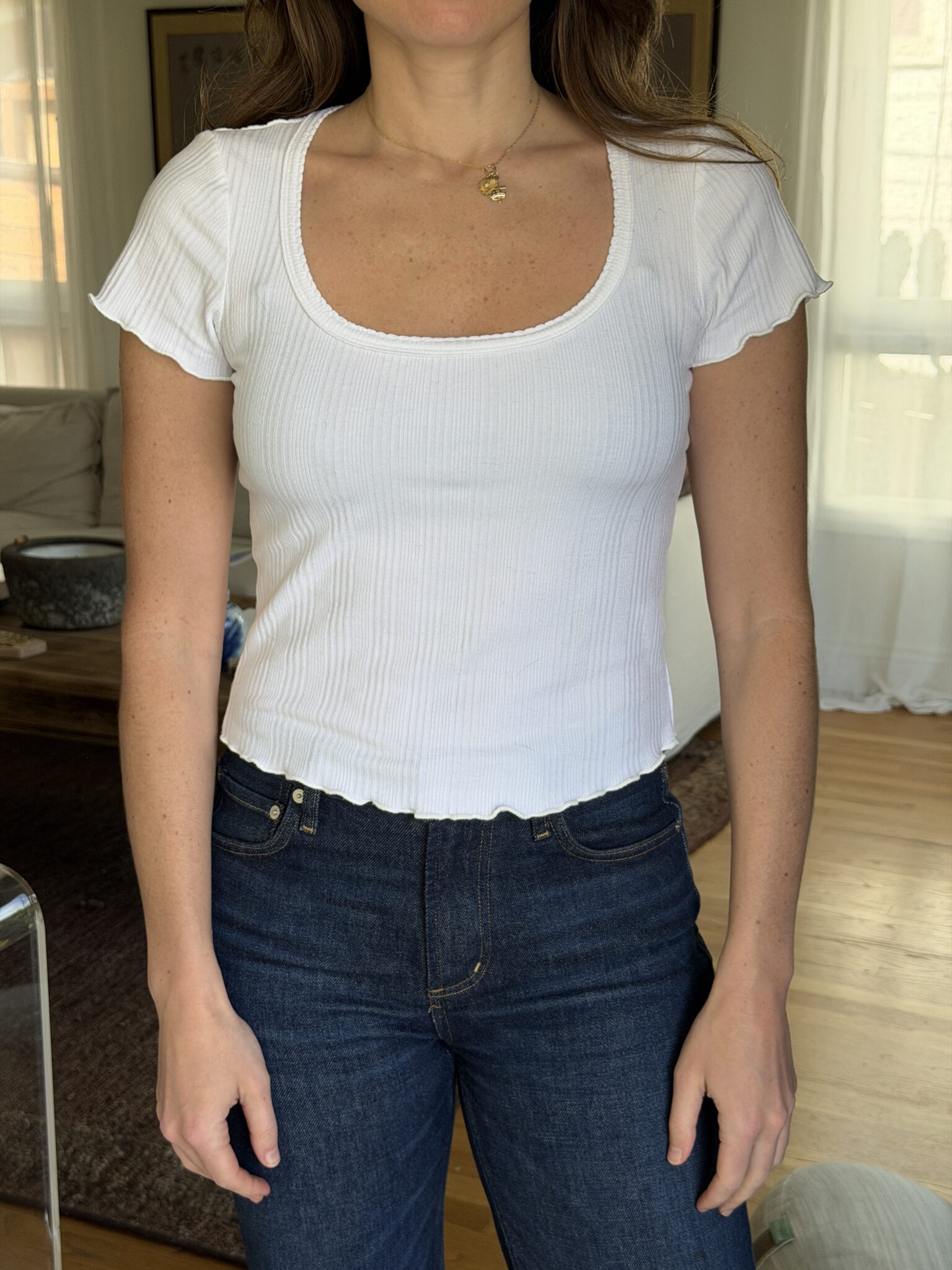 A person wearing a white short-sleeve, ribbed, scoop-neck top and dark blue jeans stands indoors with their arms relaxed at their sides.