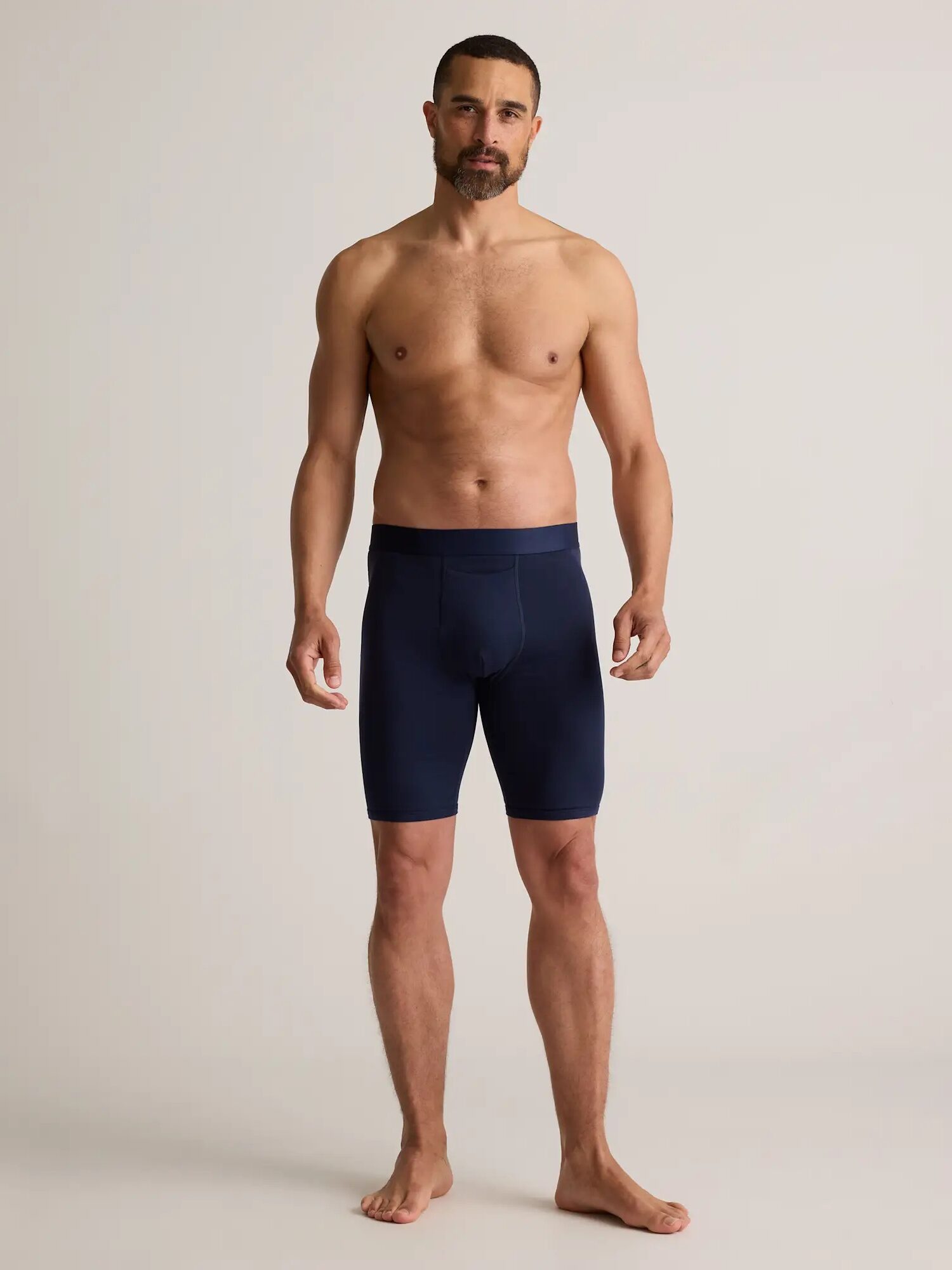 A man stands barefoot against a plain background, wearing navy blue compression shorts and no shirt.