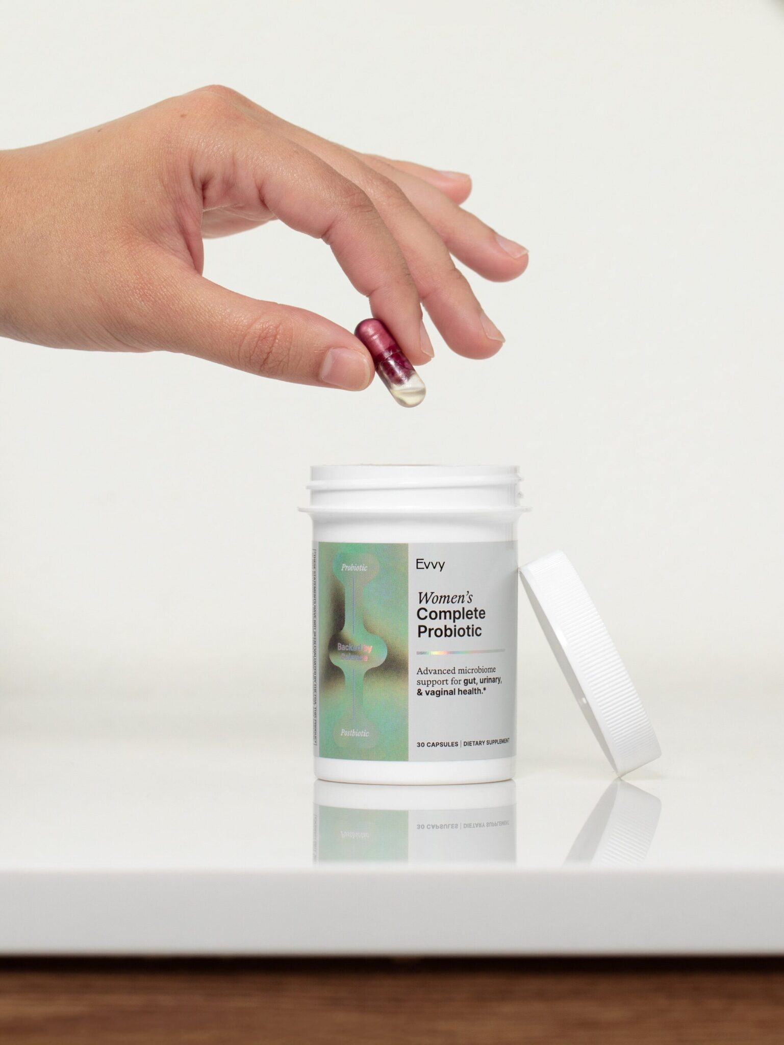 A hand holds a red capsule above an open container labeled "Women's Complete Probiotic" on a bathroom countertop.