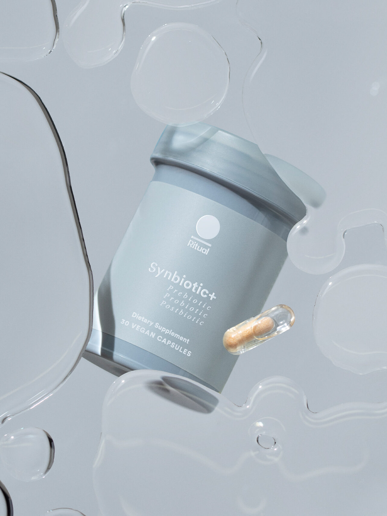 A gray container labeled "Synbiotic+ Pre & Probiotic" and a single capsule are displayed on a light gray surface with transparent liquid drops.