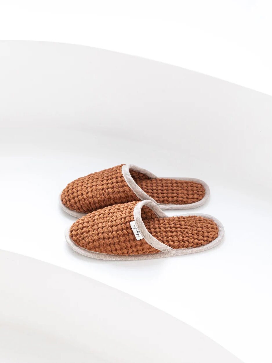 A stylish pair of brown woven slippers with white trim is placed on a white surface.