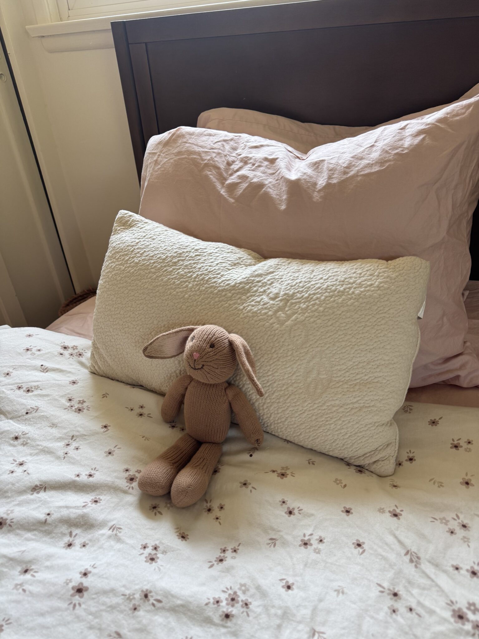 A brown stuffed rabbit sits on a white pillow, with pink bedding and floral sheets on a neatly made bed.
