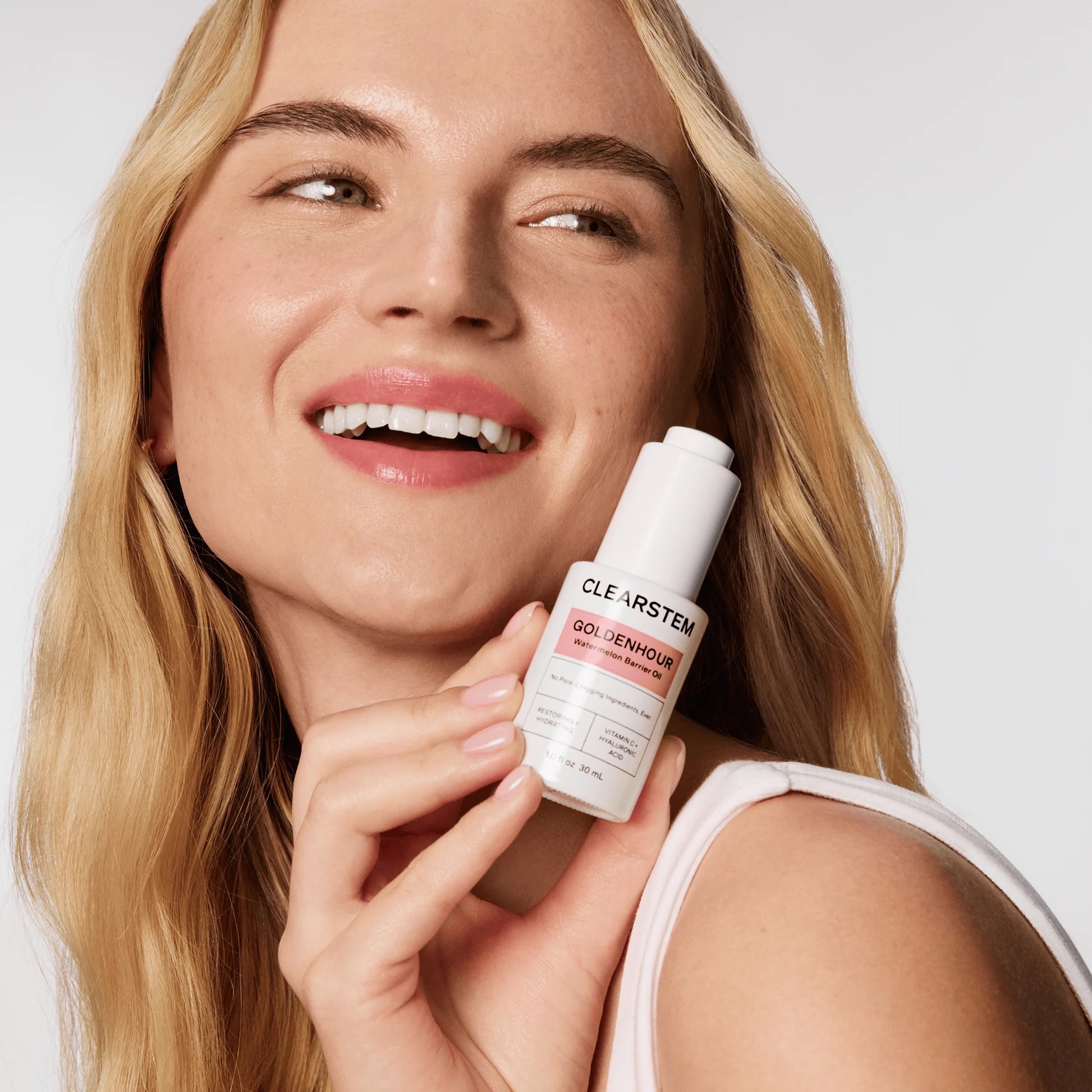 A woman with long blonde hair smiles and holds a bottle of CLEARSTEM GOLDEN HOUR skincare serum close to her face against a plain background.