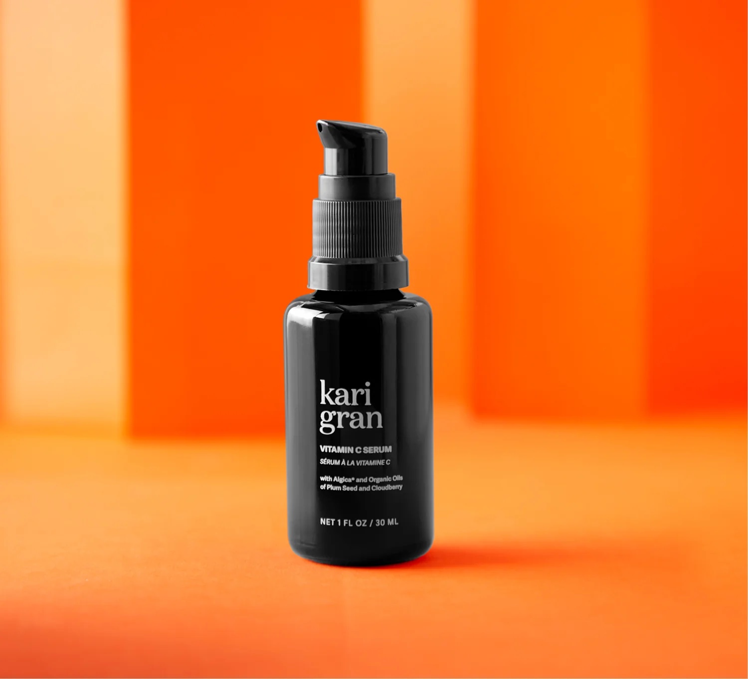 A black pump bottle labeled "Kari Gran Vitamin C Serum" sits on an orange surface with an orange gradient background.