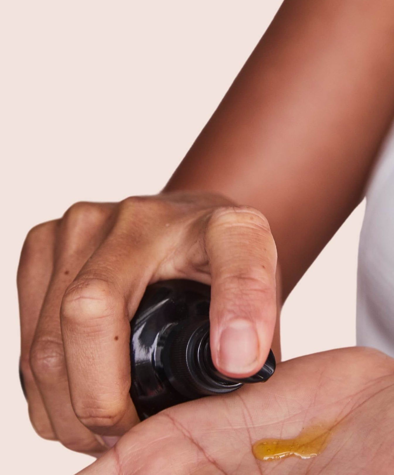 A person dispenses a golden liquid from a dark bottle onto their palm against a plain, light background.
