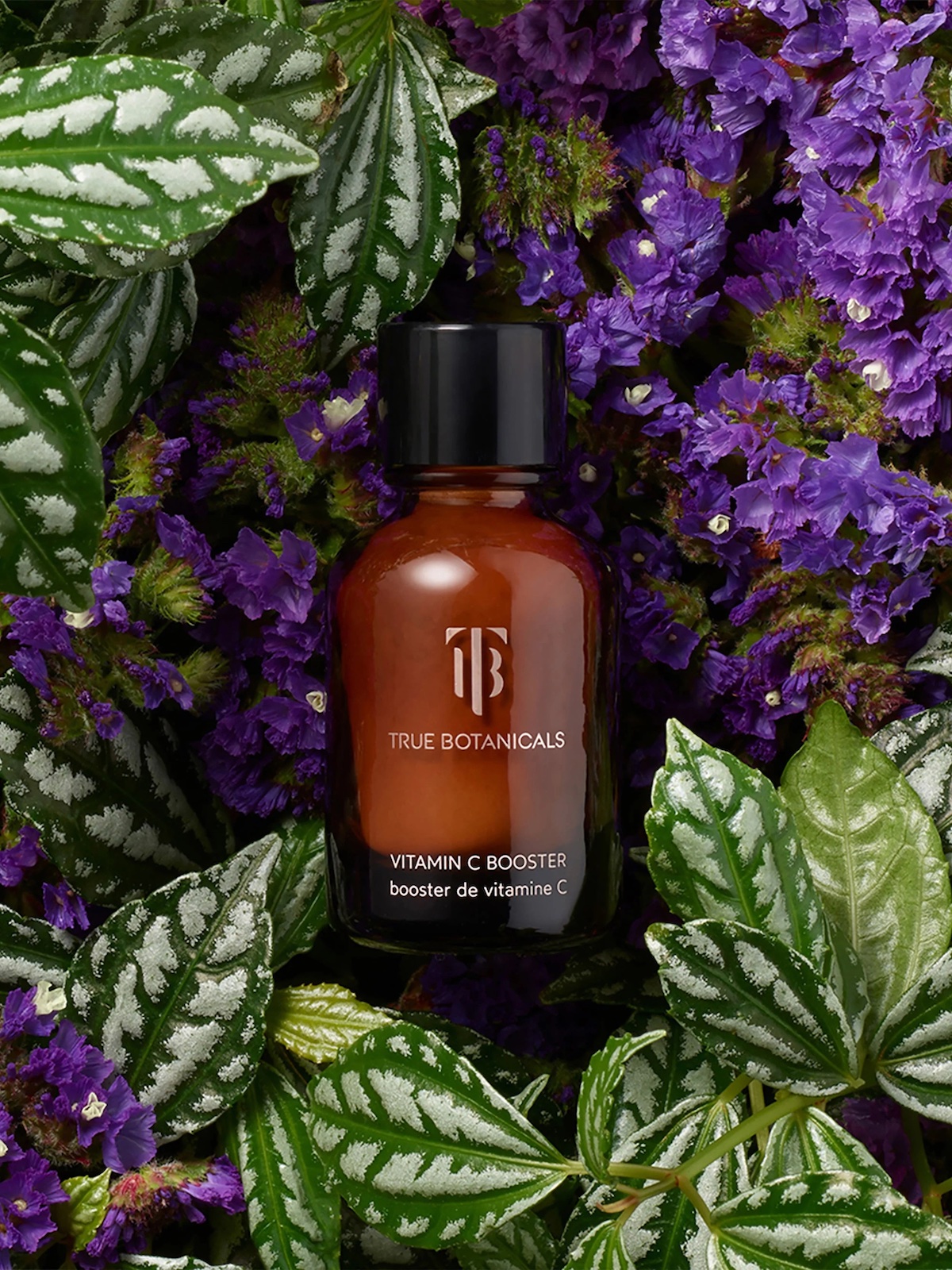 A brown glass bottle labeled "True Botanicals Vitamin C Booster" is surrounded by purple flowers and green leaves.