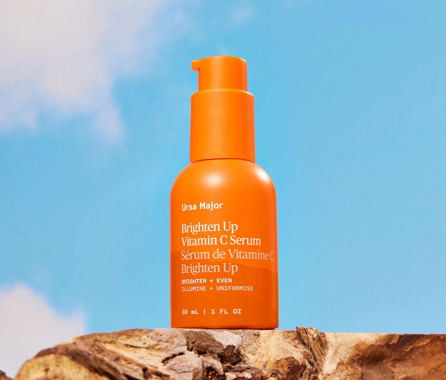 An orange bottle of Ursa Major Brighten Up Vitamin C Serum sits on a rock against a bright blue sky with clouds.