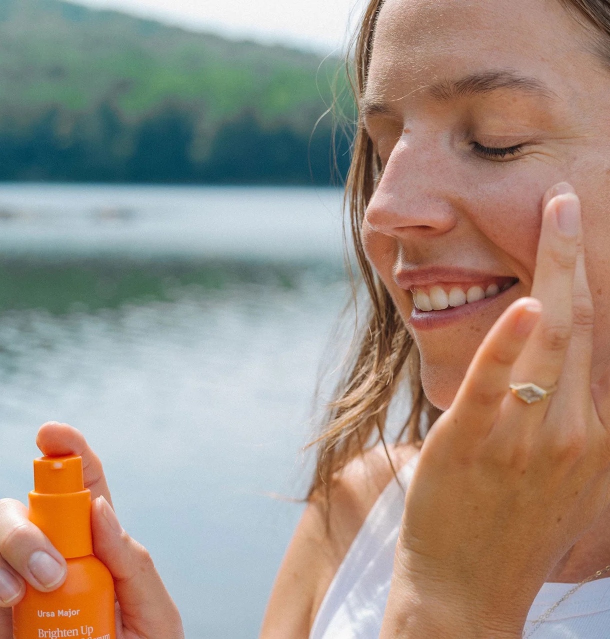 A woman applies skincare serum to her face while standing outdoors near a lake, holding an orange bottle labeled "Ursa Major Brighten Up.