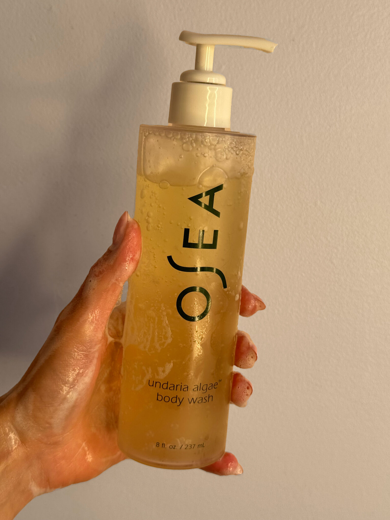 A hand holding a clear pump bottle of OSEA undaria algae body wash against a plain light-colored background.