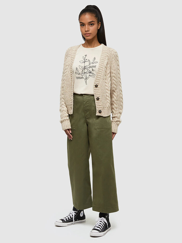 A woman stands wearing a cream cable knit cardigan, a white shirt with a botanical print, olive green wide-leg pants, and black high-top sneakers.