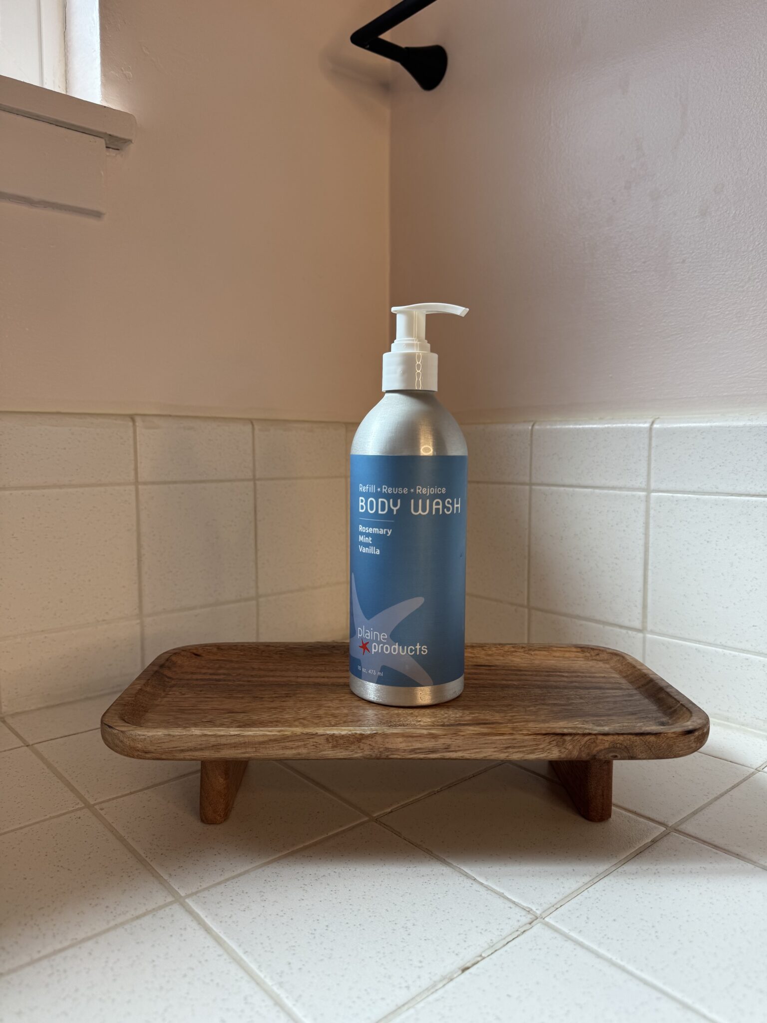 A bottle of body wash with a pump sits on a small wooden tray in the corner of a tiled bathroom countertop.