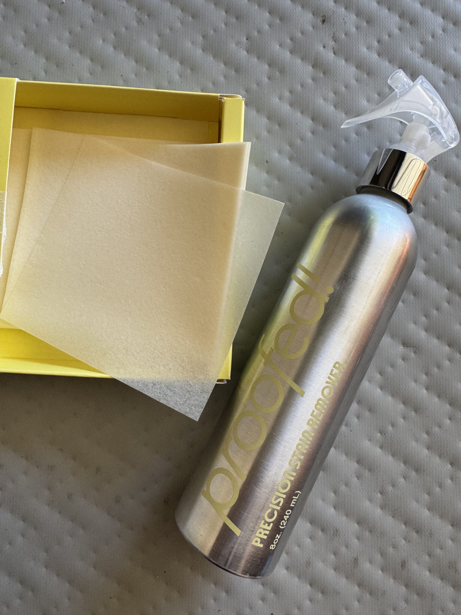 A spray bottle labeled "proofed! PRECISION SHRINKPROOF" next to a yellow box containing thin translucent sheets, on a textured surface.
