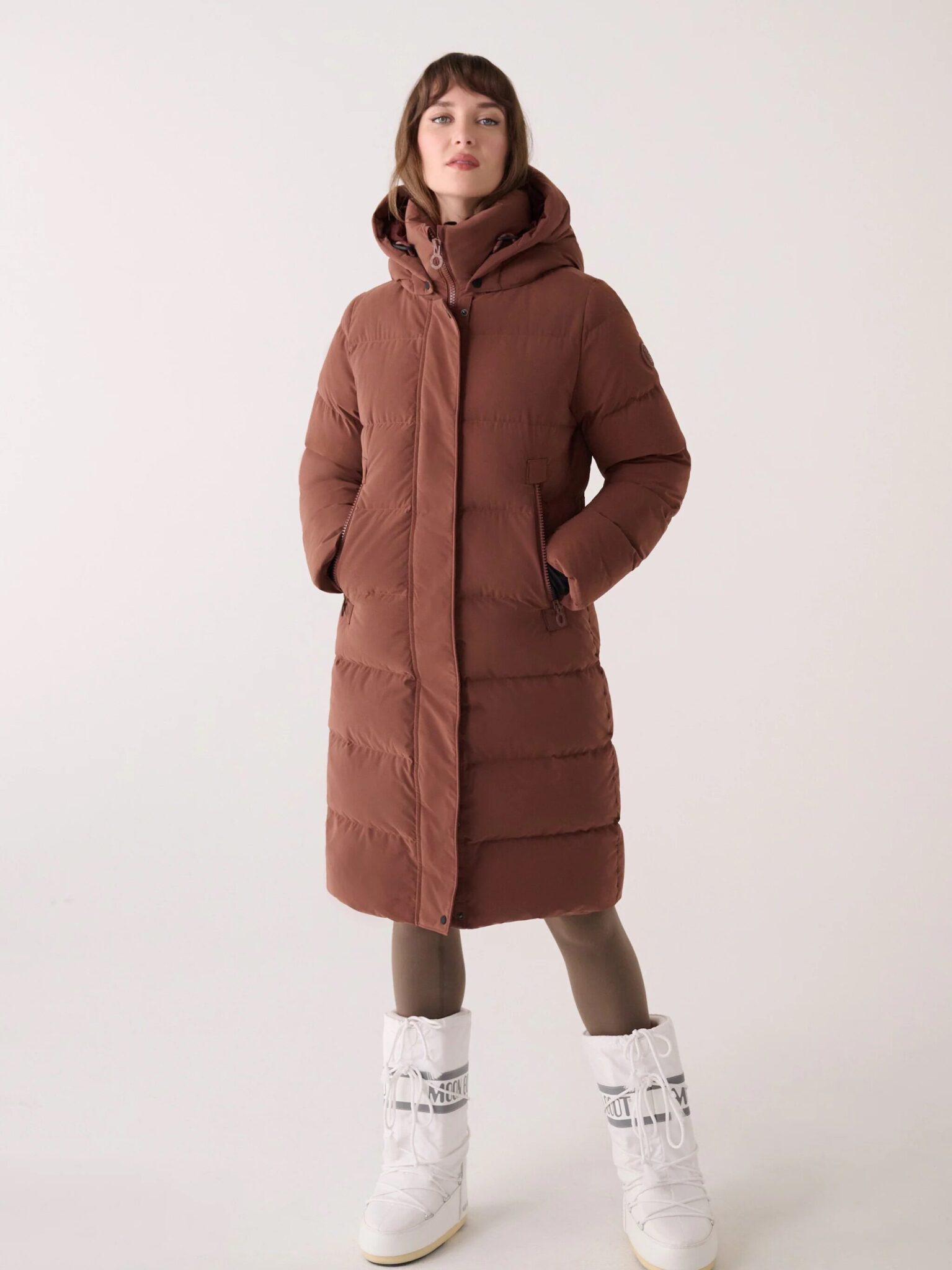 A person wearing a long brown puffer coat, brown leggings, and white snow boots stands against a plain light background.