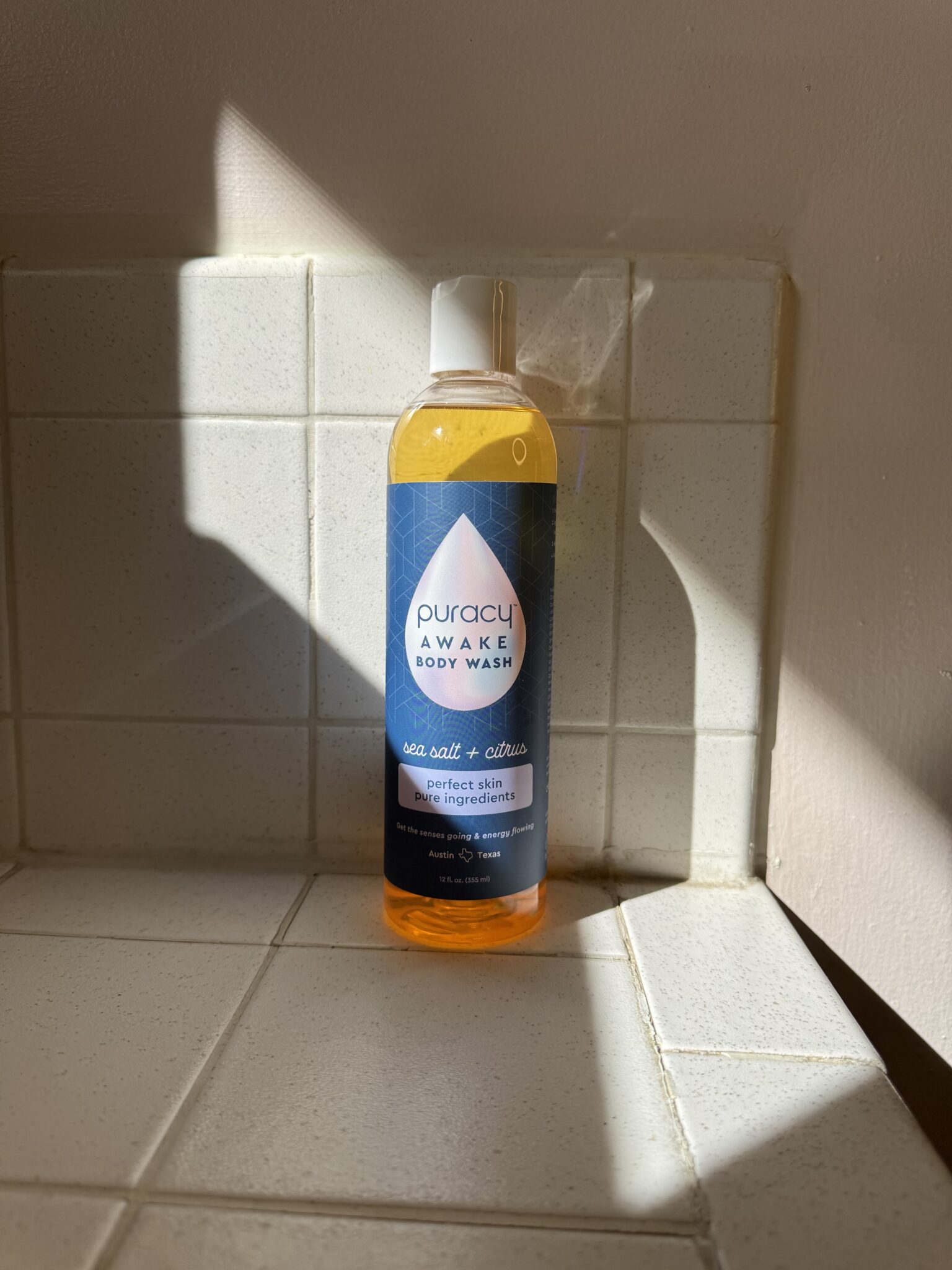 A bottle of Puracy Awake Body Wash with sea salt and citrus stands on a tiled bathroom surface in sunlight.