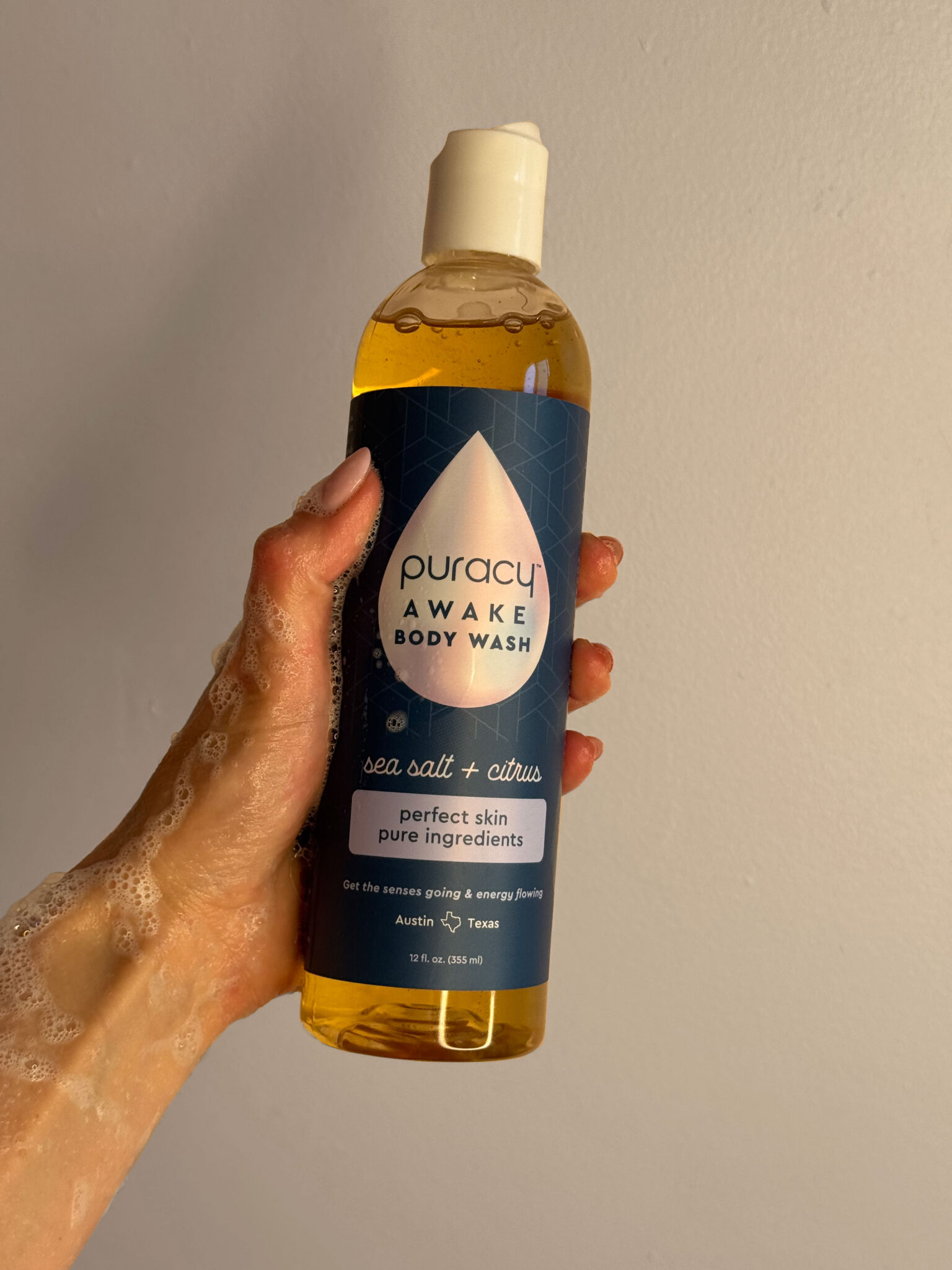 A hand holding a bottle of Puracy Awake Body Wash, sea salt and citrus scent, against a plain beige background.