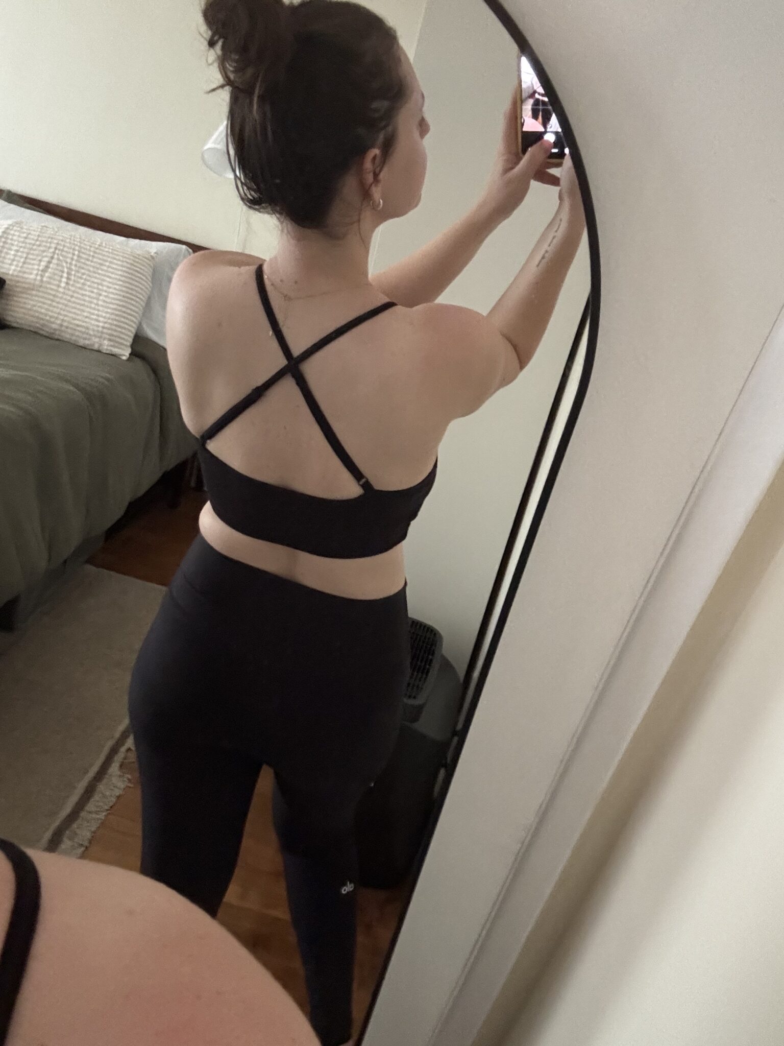 Person taking a mirror selfie showing the back of their black sports bra and leggings in a bedroom with a made bed in the background.