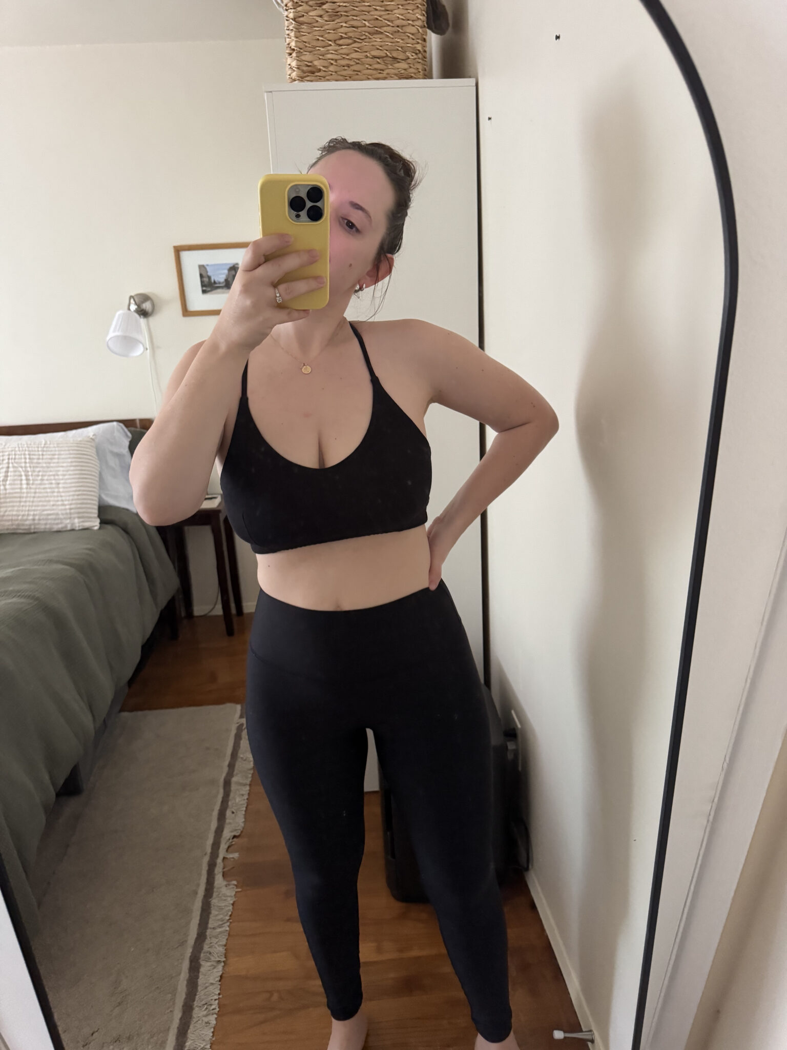 A person wearing a black sports bra and leggings takes a mirror selfie in a bedroom with a neatly made bed and simple decor.