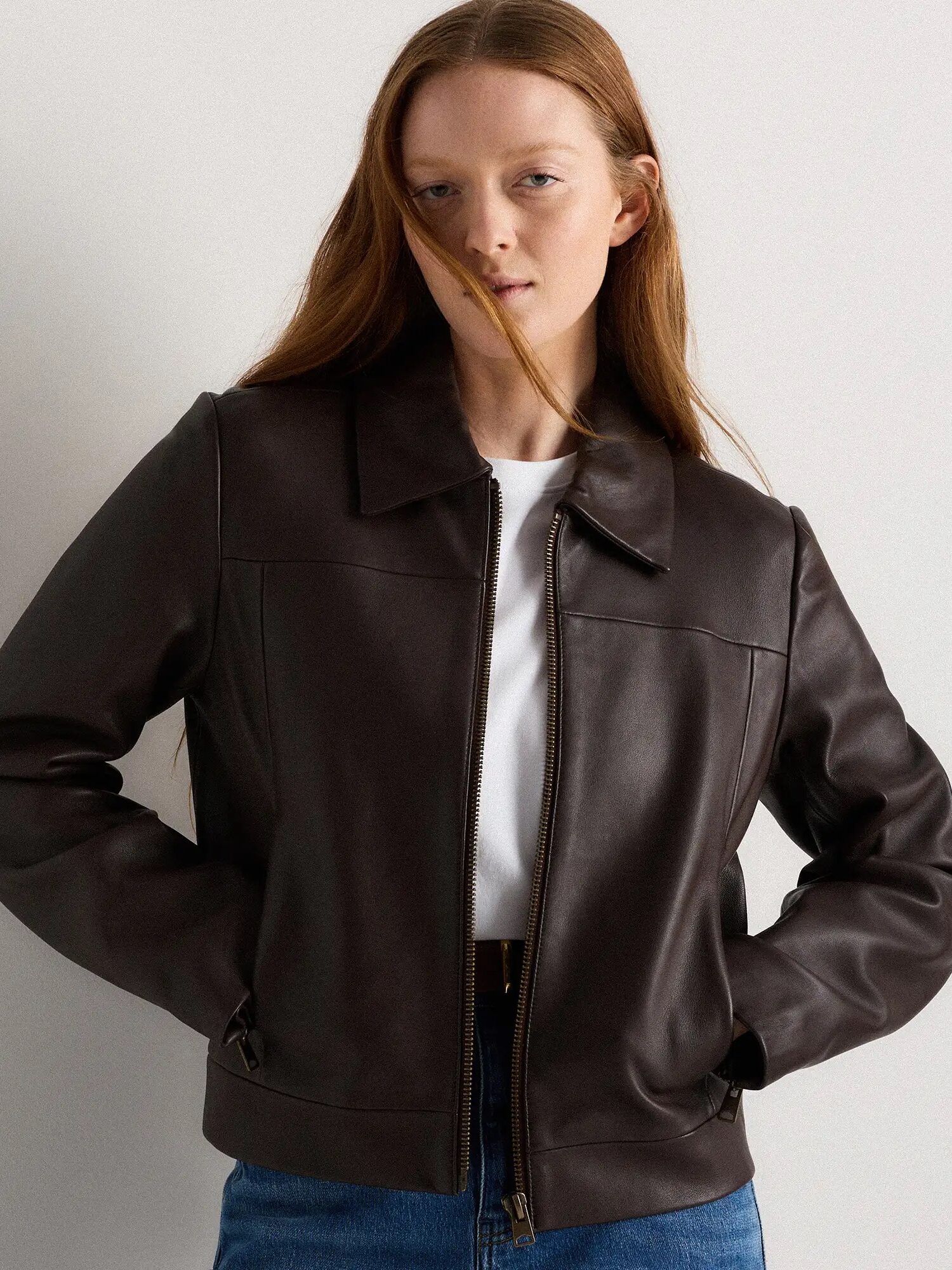 A person with long hair wears a dark brown leather jacket over a white shirt and blue jeans, standing against a plain light background.