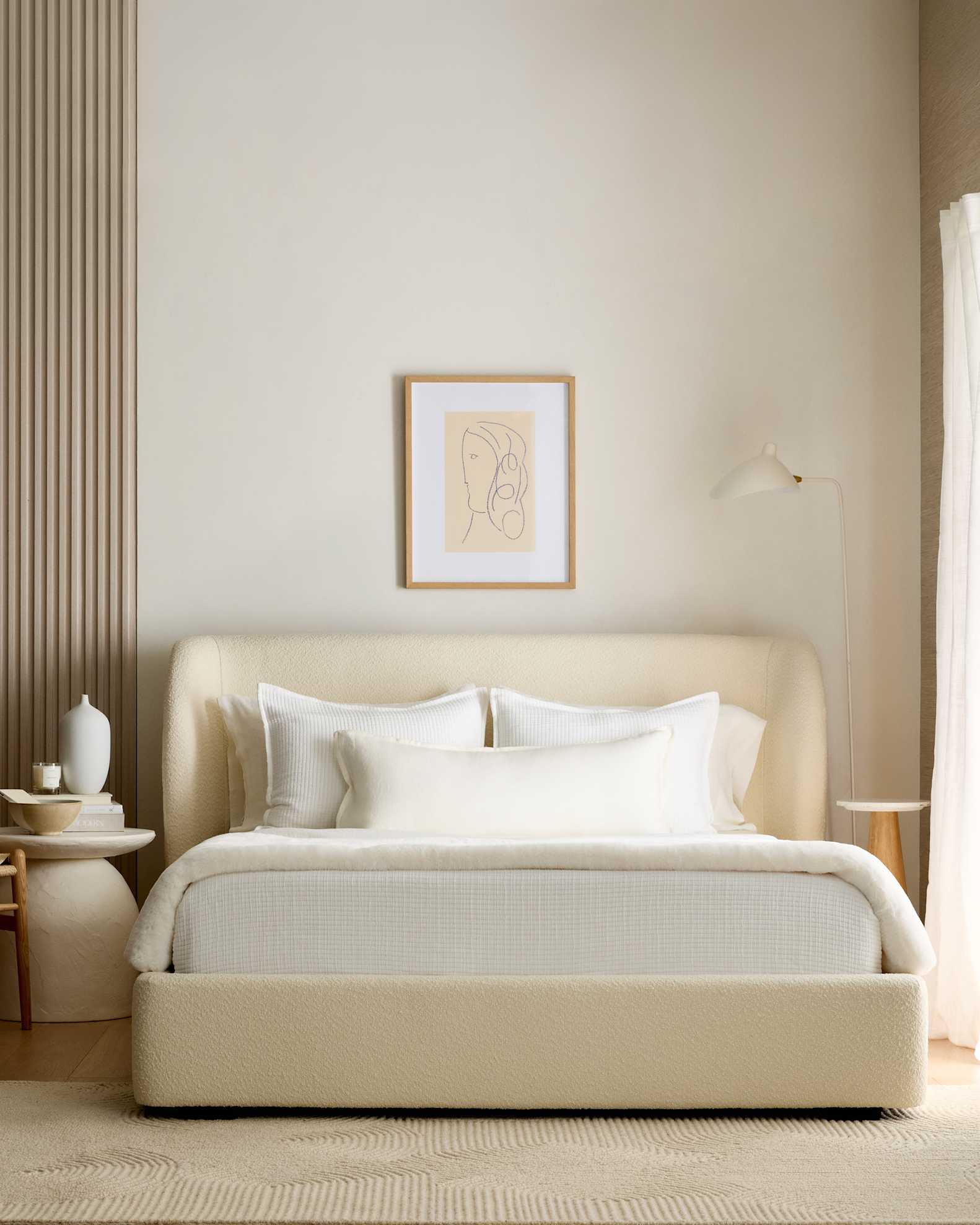 A minimalist bedroom features a cream bed with white linens, a framed abstract line drawing above the headboard, and light-colored decor on side tables.